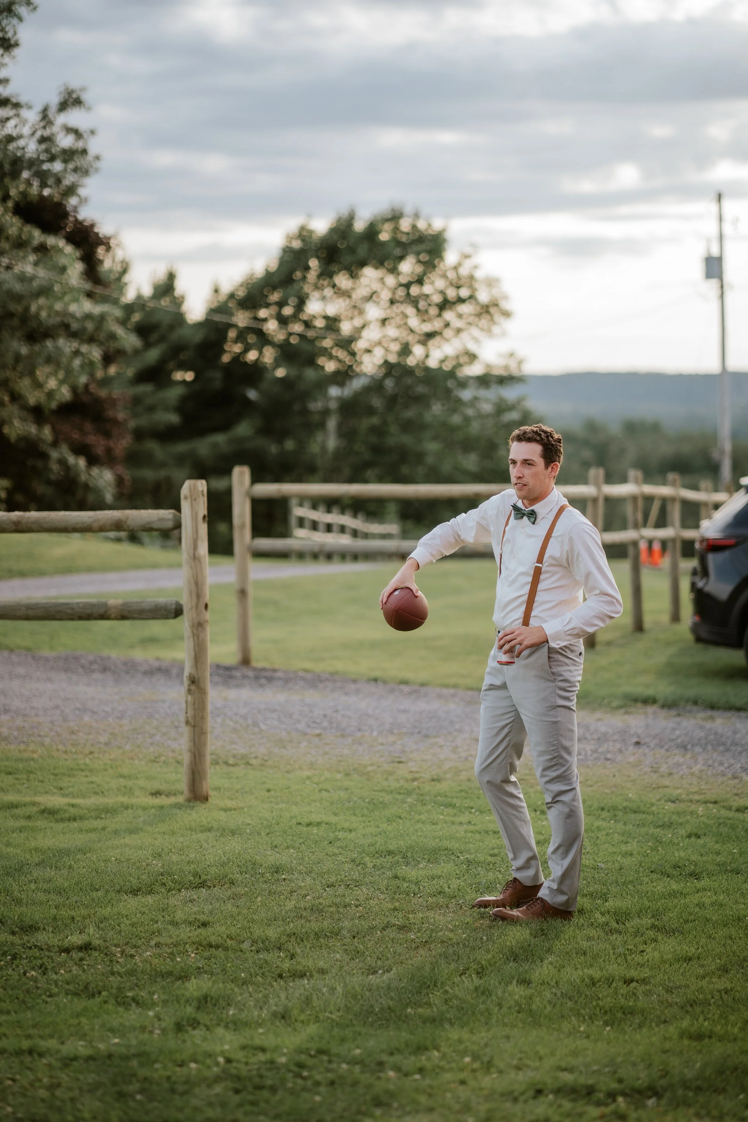 A man in formal attire, wearing a white shirt, light gray pants, suspenders, and a bow tie, stands on a grassy area holding an American football in his right hand and a drink in his left hand during late afternoon or early evening.