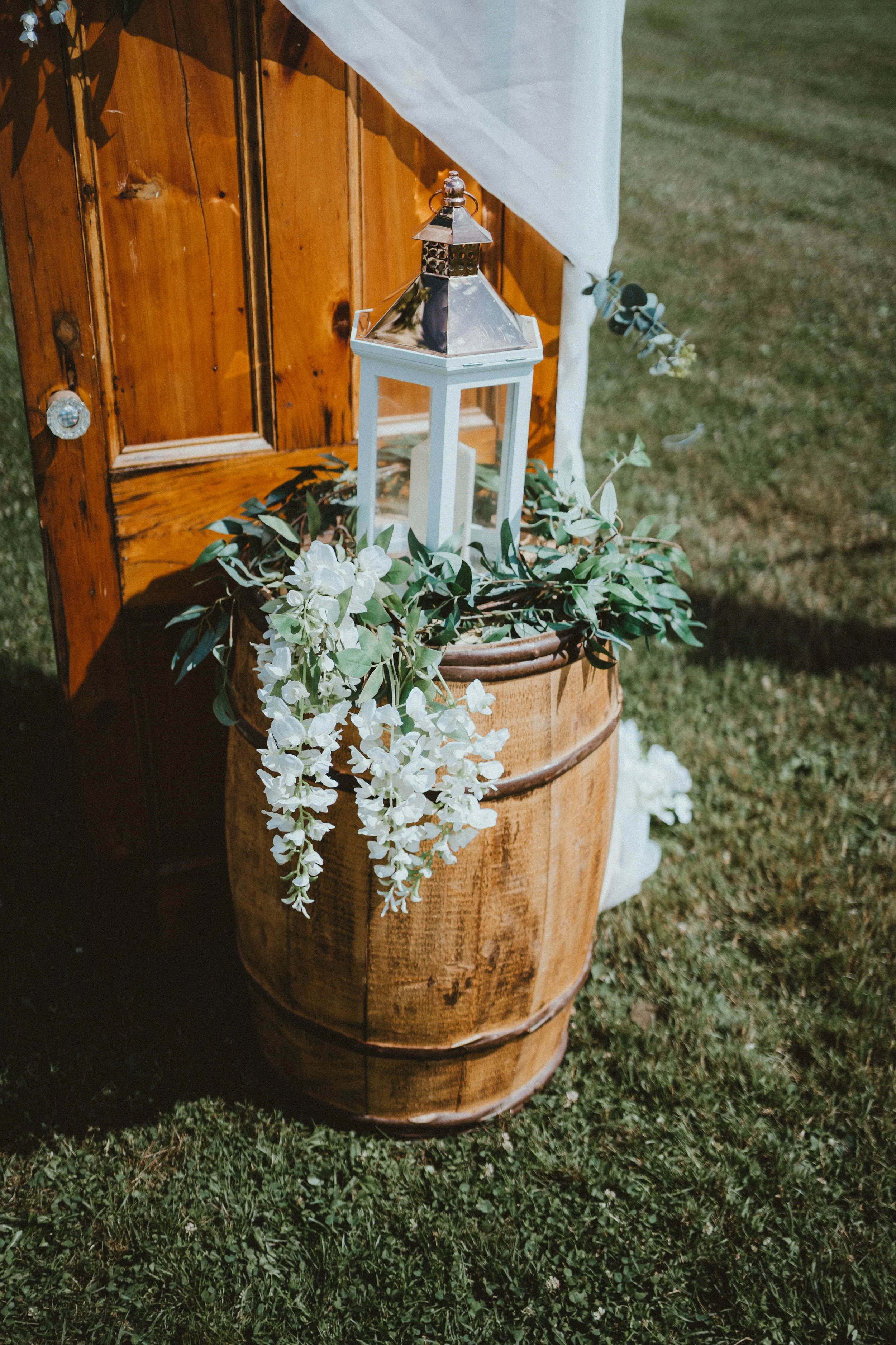 A decorative outdoor setup with a white lantern on top of a wooden barrel, adorned with white flowers and green foliage, next to a wooden structure with white fabric draping.