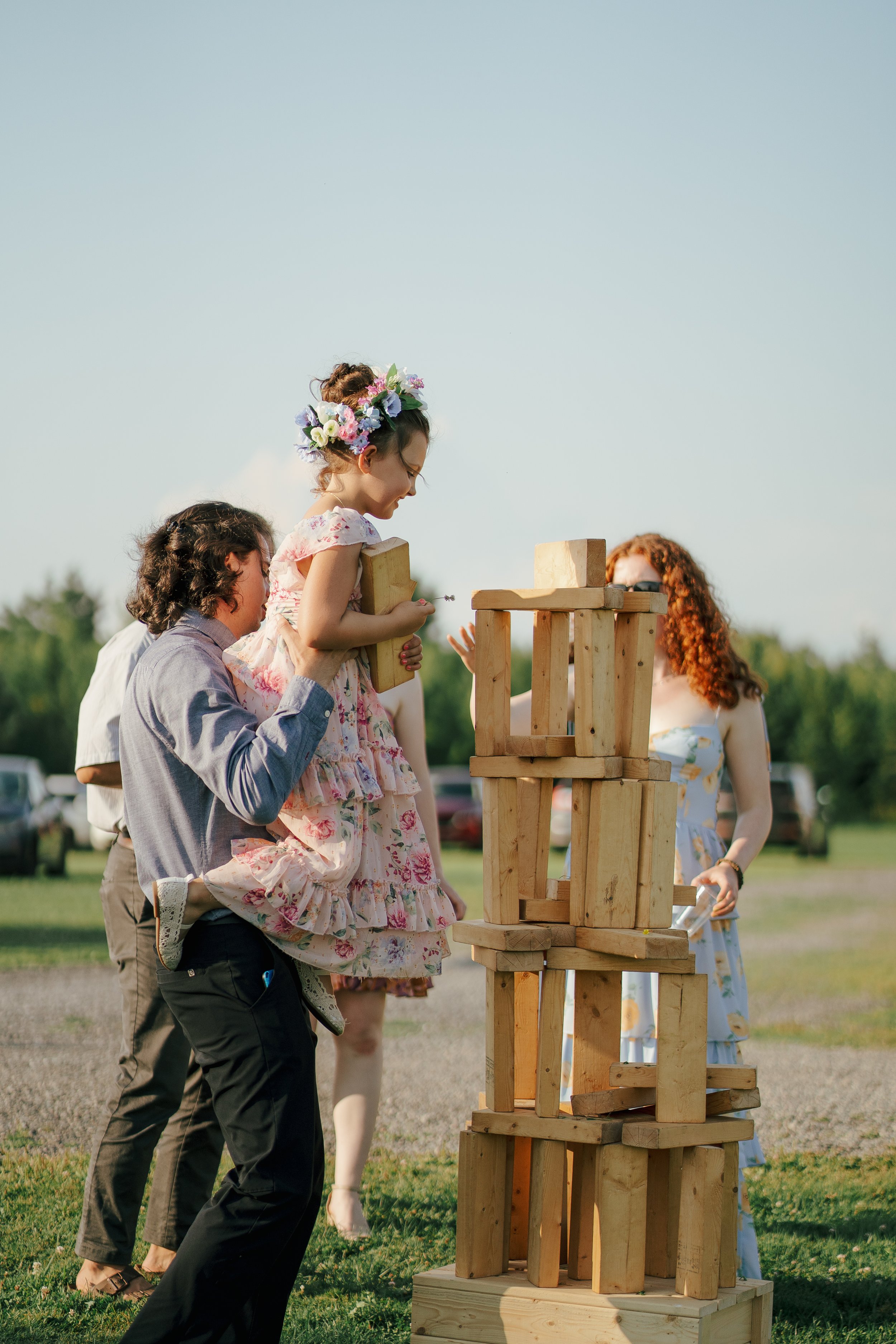 Children and adults playing a giant Jenga game outdoors on a grassy field.