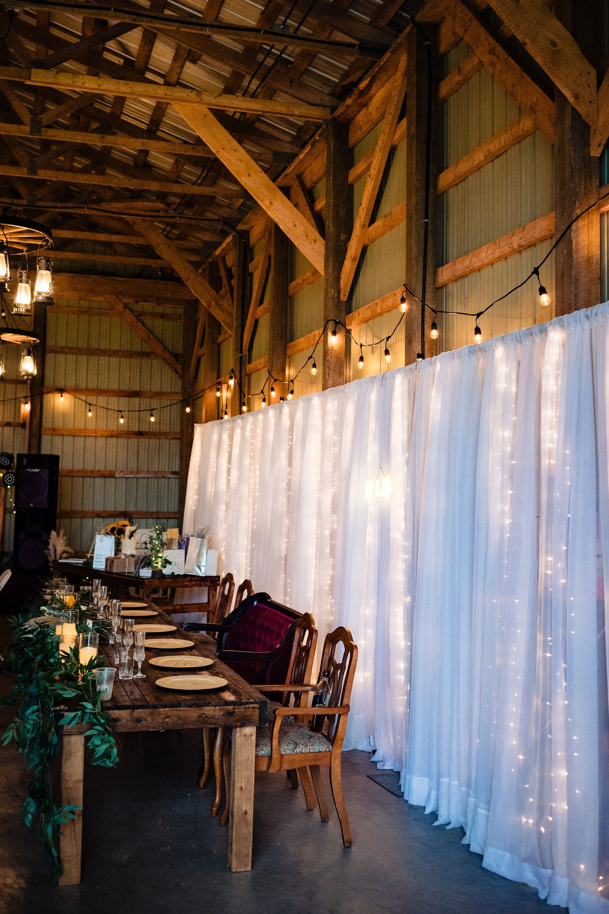 A decorated event space with a rustic wood interior, string lights, a white curtain backdrop with fairy lights, and a long table set for a celebration.