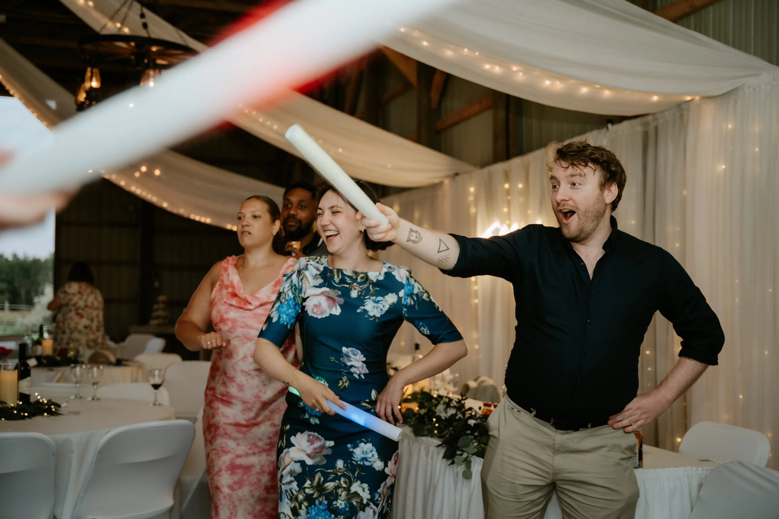 People at a celebration, including a man in a black shirt holding a glow stick, a woman in a blue floral dress, and others, inside a decorated barn with string lights.