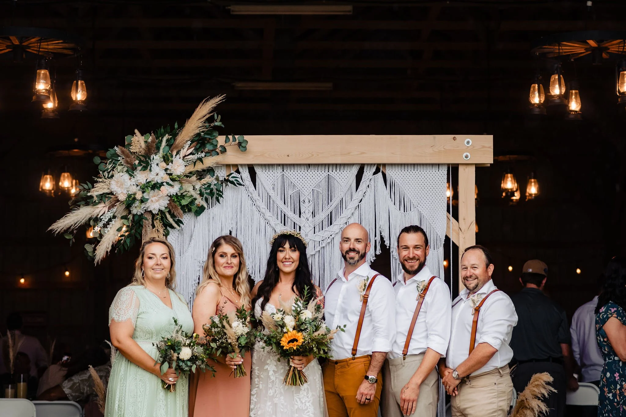 Group of wedding guests standing together in front of a rustic backdrop decorated with flowers and macrame at an indoor wedding reception.