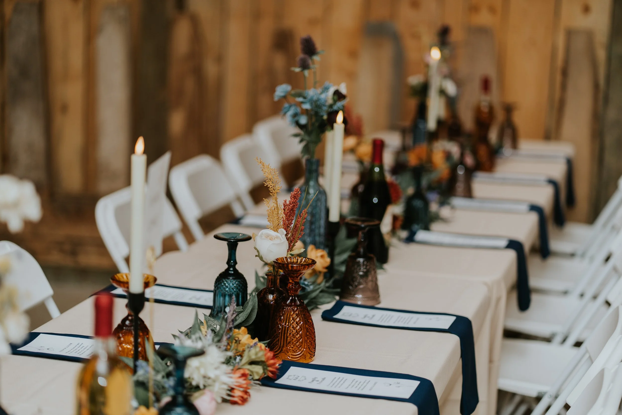 Decorated banquet table with colorful vases, flowers, tall candles, menus, and white chairs in a rustic wooden venue.
