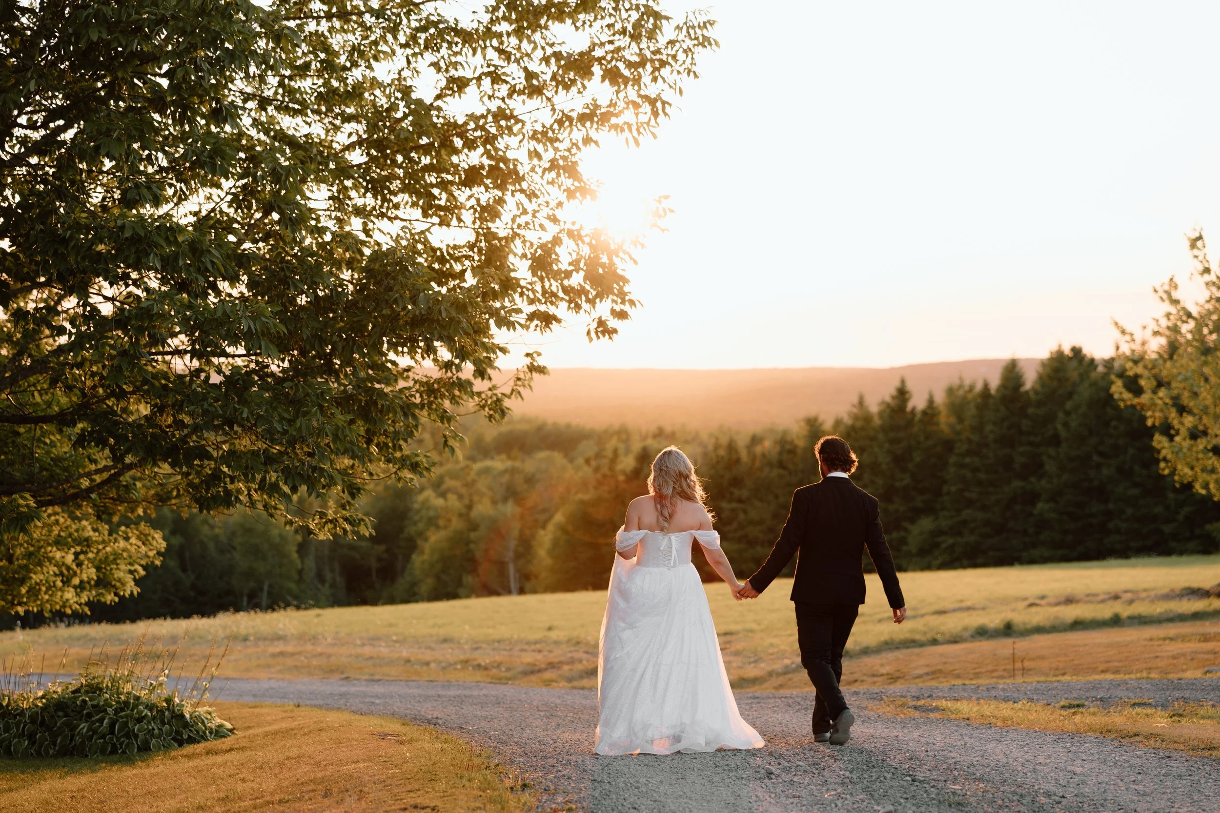 A bride and groom walking hand in hand on a gravel path during sunset in a rural landscape with trees and open fields.