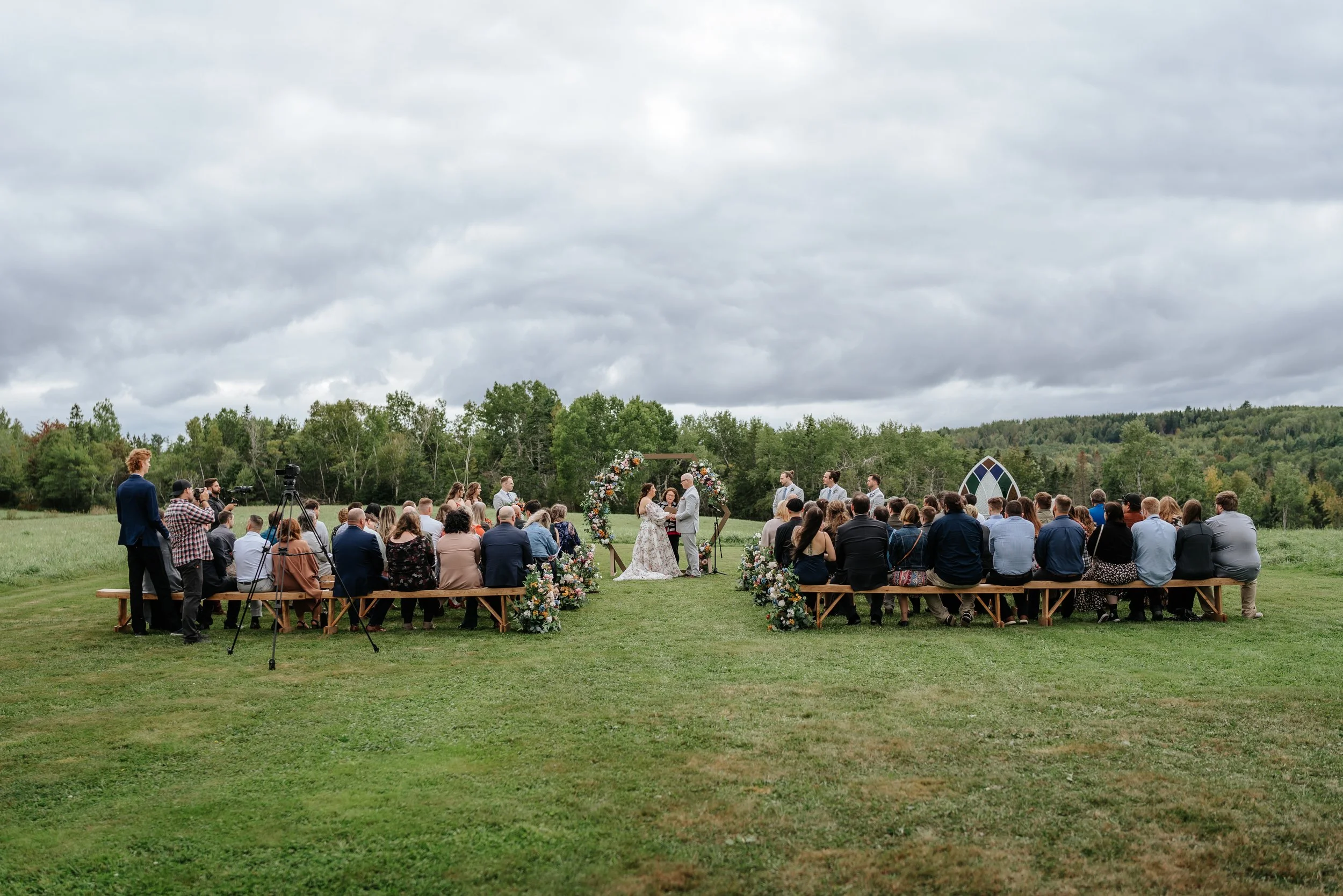 Outdoor wedding ceremony set on a grassy field with a large group of guests sitting on benches, facing a couple exchanging vows under a floral arch with a scenic backdrop of trees and cloudy sky.