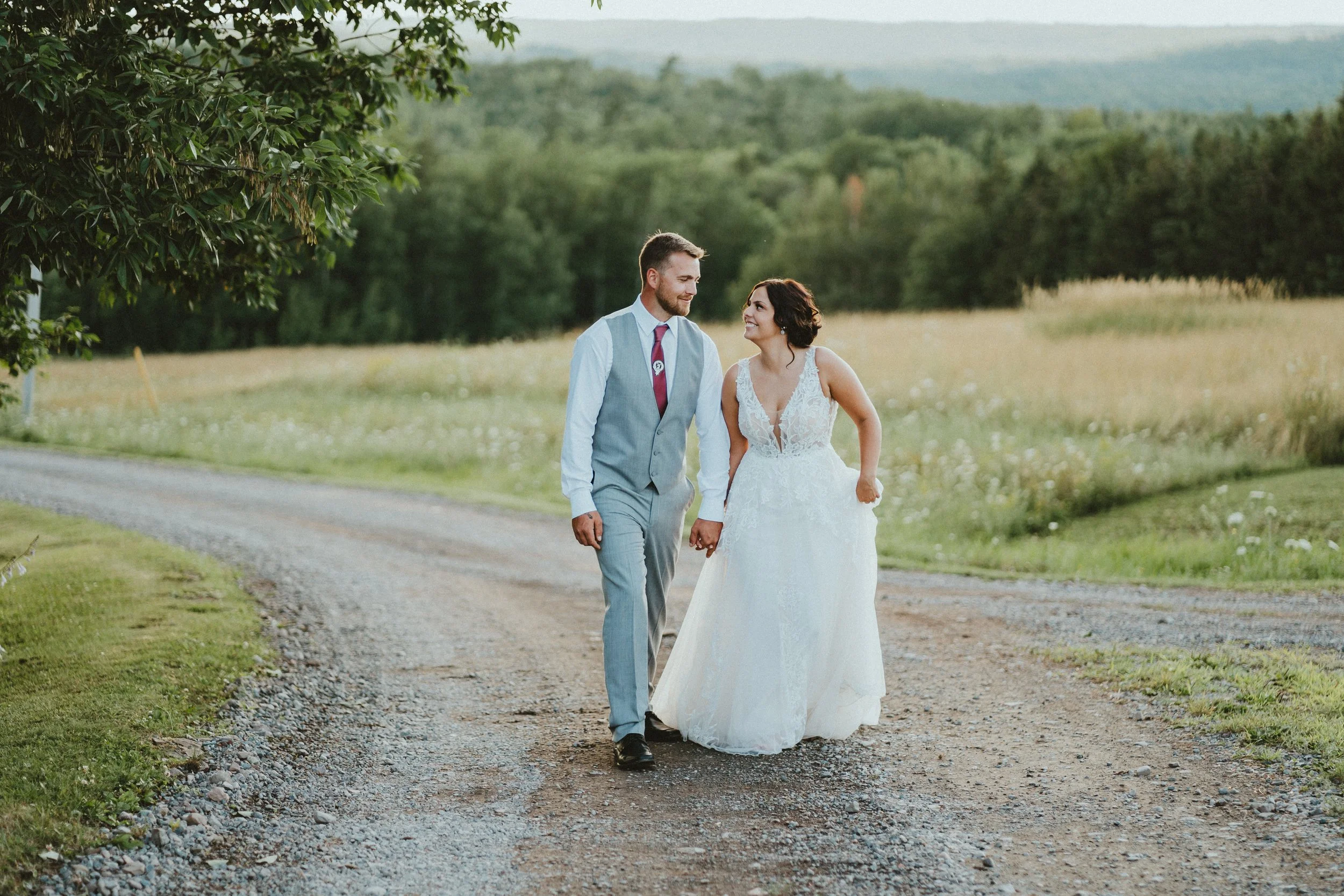 A bride and groom walking hand in hand on a dirt path in a rural setting, smiling at each other.