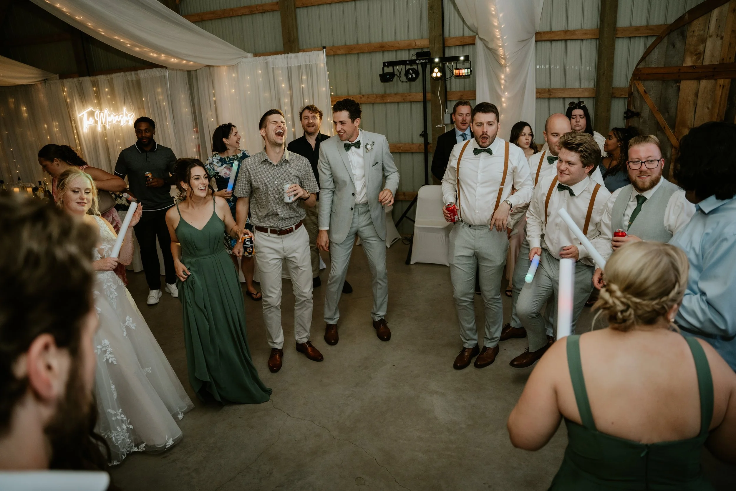 Wedding reception with guests dancing in a barn. The group is smiling and enjoying themselves, some holding glow sticks and drinks, with wedding decorations and lights in the background.