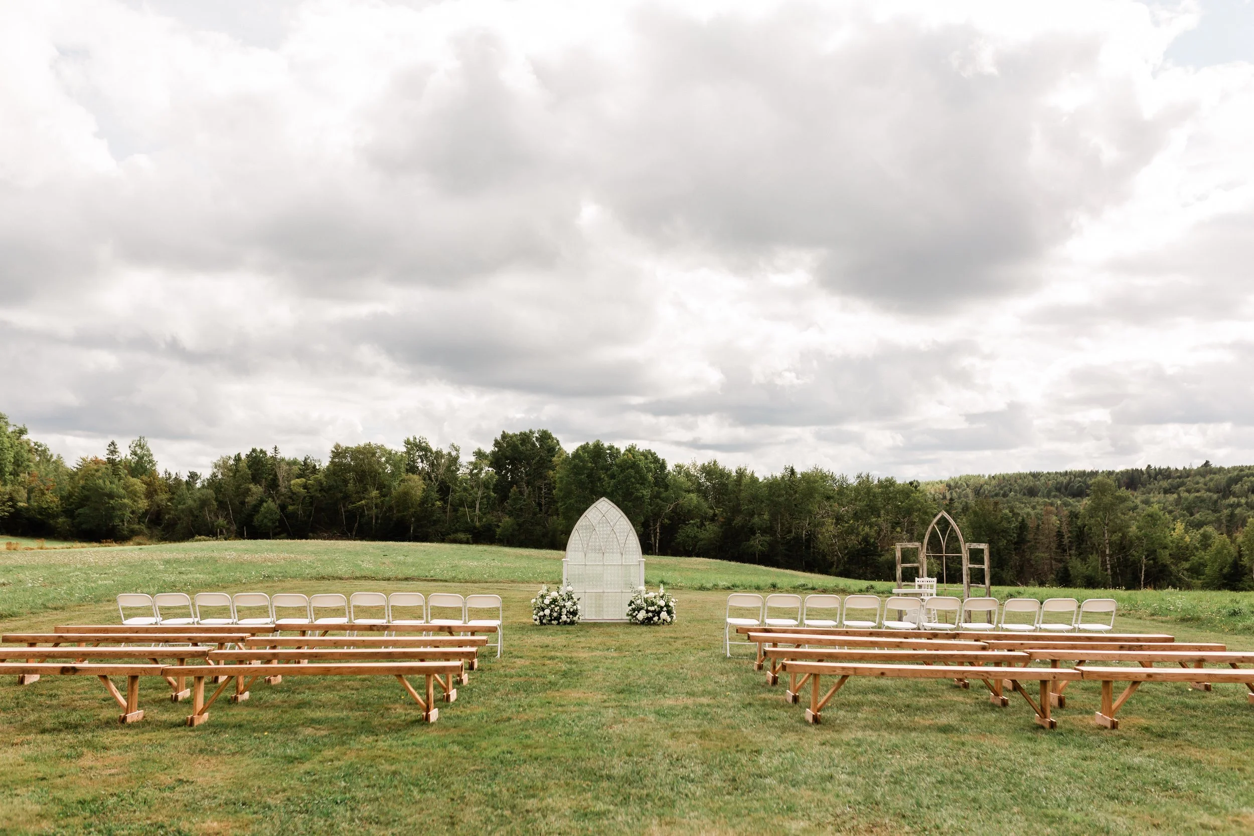 Outdoor wedding setup with wooden benches and white chairs facing a floral arch and a small table on a grassy field, with trees and cloudy sky in the background.