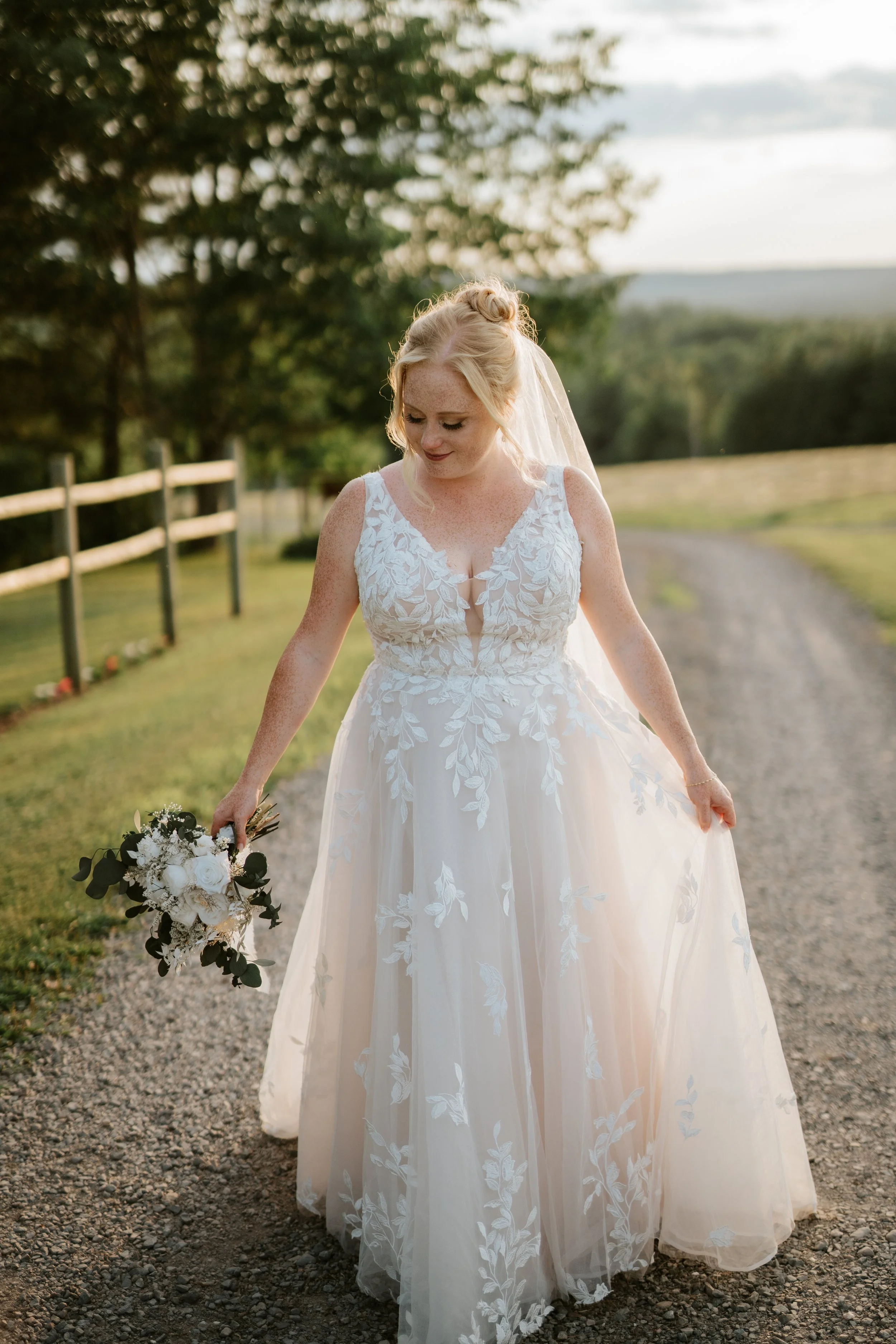 A bride in a white lace wedding dress holding a bouquet of white flowers and greenery, walking outdoors on a gravel path during sunset.