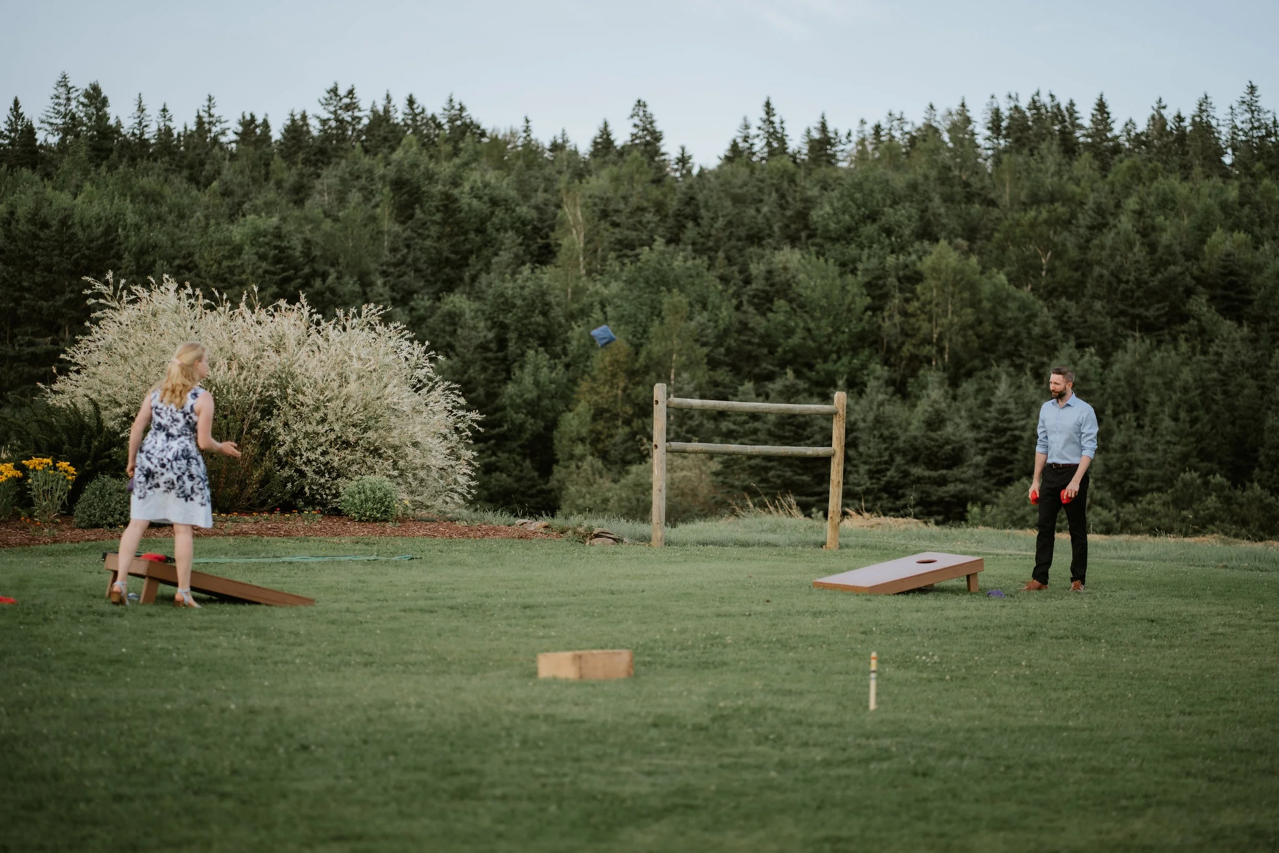 A man and a woman playing cornhole outdoors on a grassy field with trees in the background.