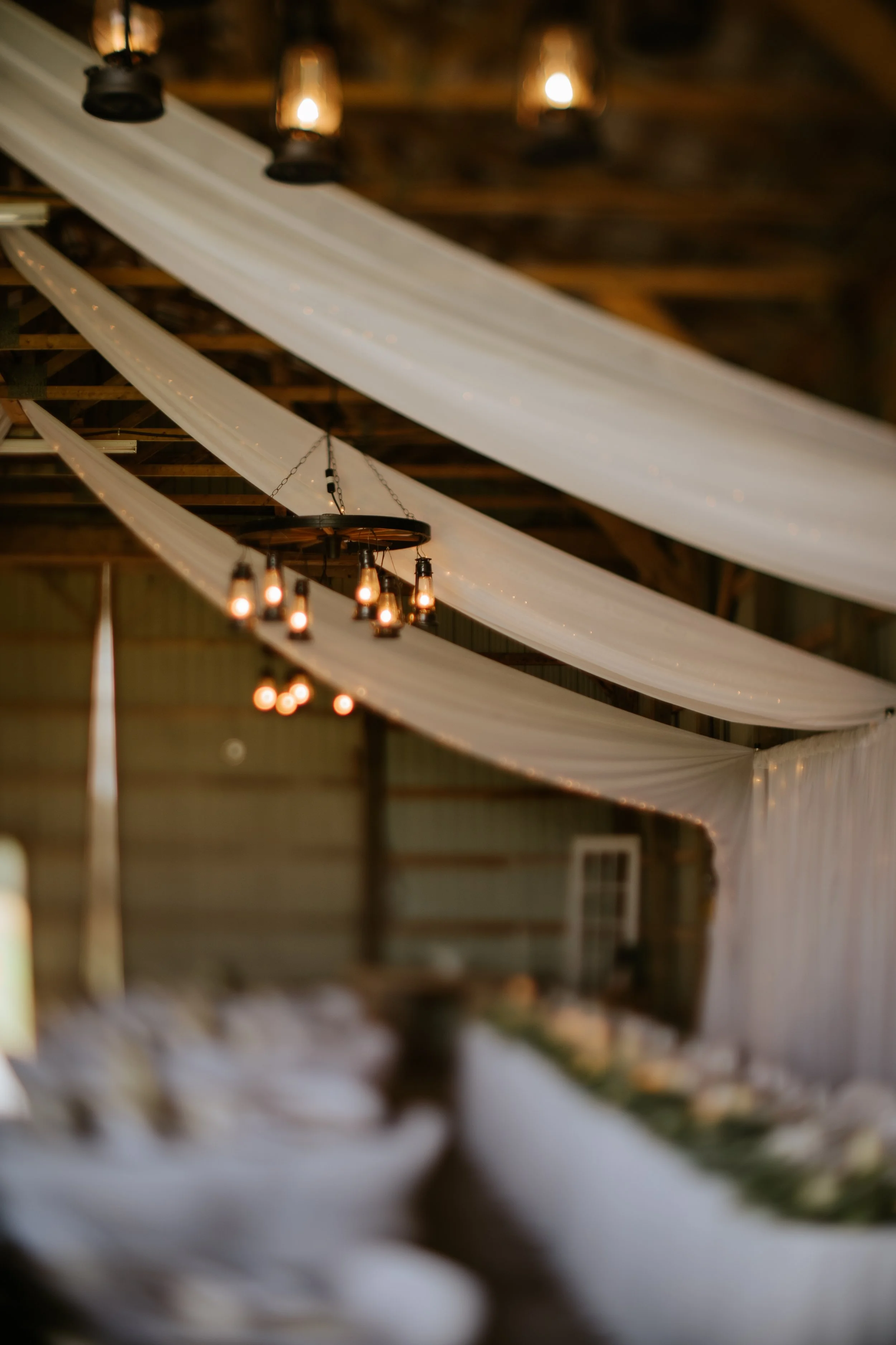 Decorated wedding reception area featuring white drapery hanging from the ceiling with warm string and chandelier lights.
