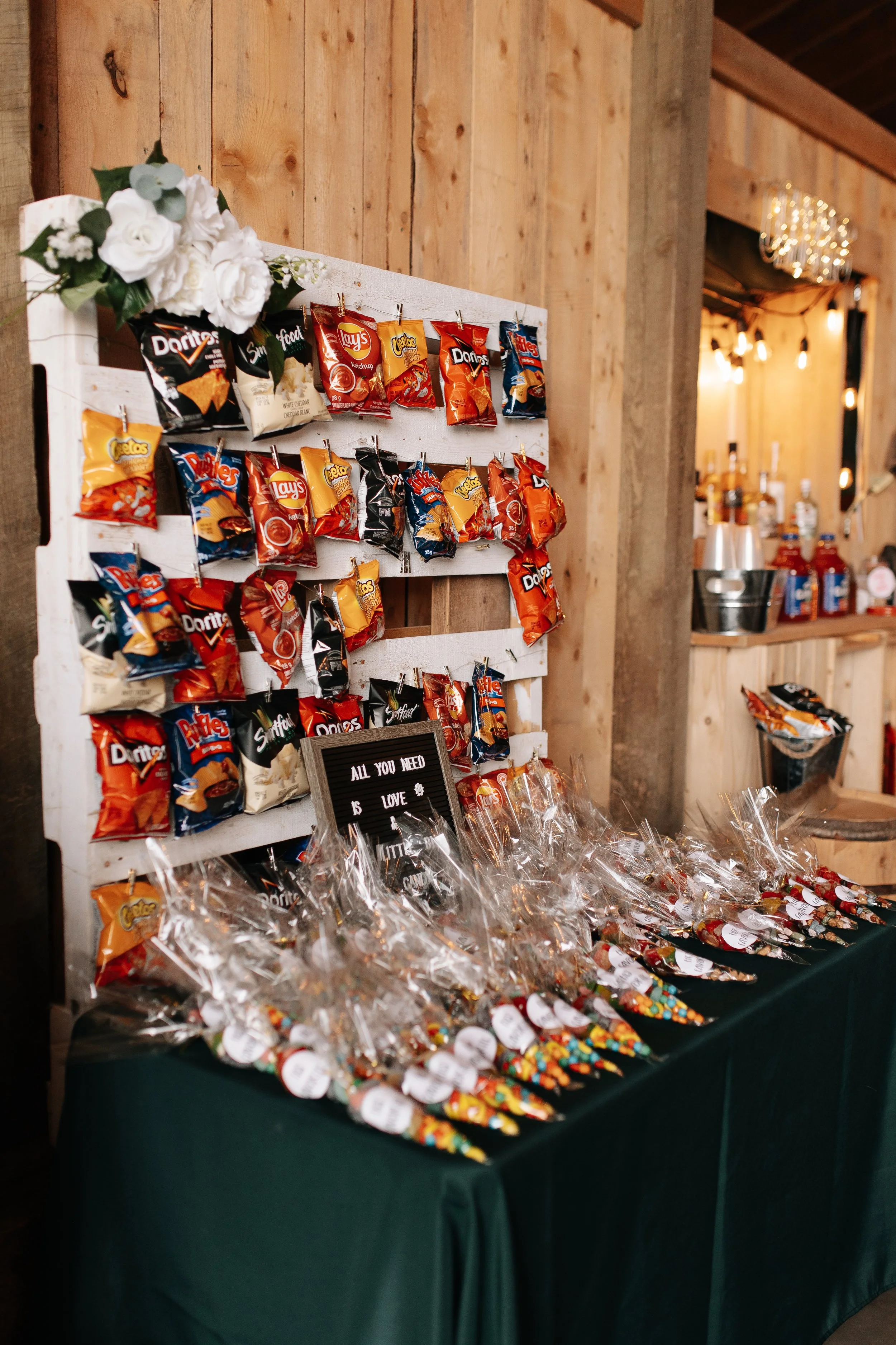 A display of assorted snack bags and gift bags on a table at a party or event with a decorative wooden backdrop and string lights.