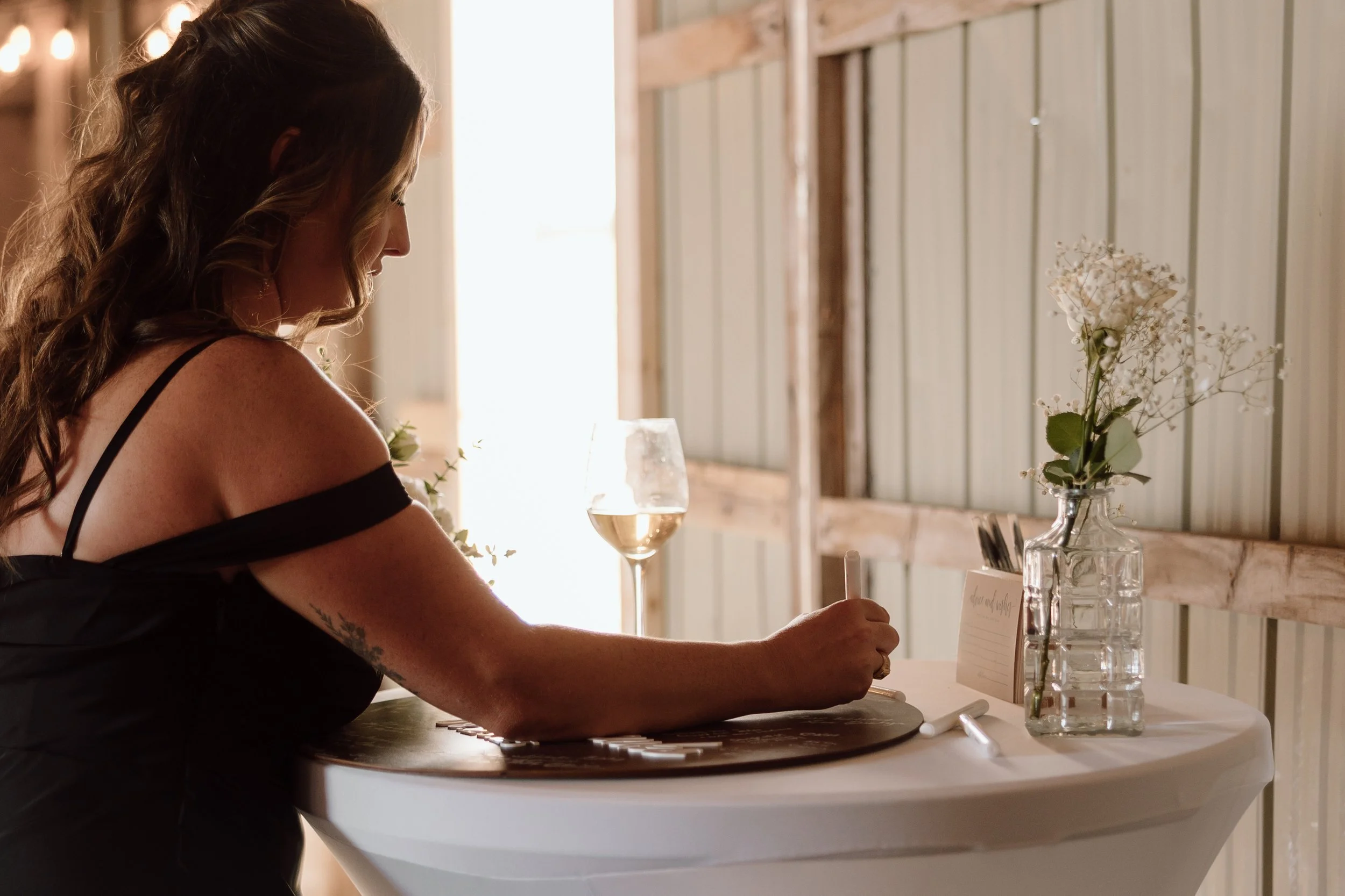 A woman with long wavy hair, wearing a black dress, sitting at a table and writing on a card with a pen, in a rustic setting with a glass of white wine and a vase with white flowers.