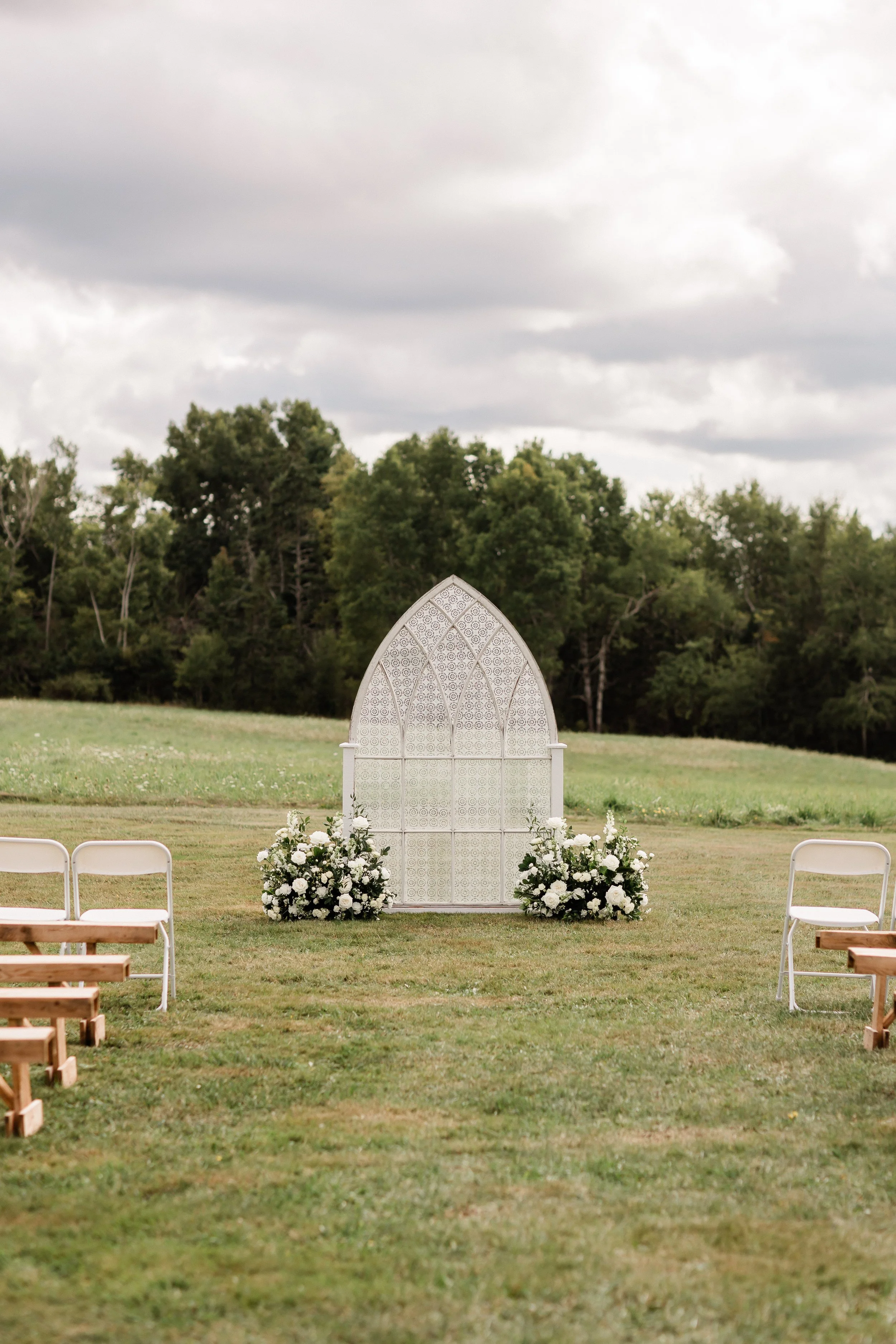 Outdoor wedding ceremony setup with a white decorative arch adorned with white flowers, green foliage, set on a grassy field surrounded by trees. White chairs are arranged on either side of the aisle under cloudy sky.