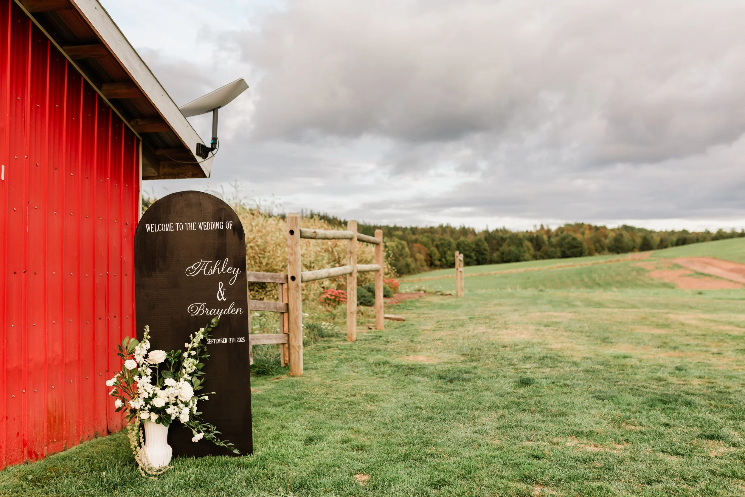 Outdoor wedding setup with a welcome sign for Ashley and Brayden, along with a vase of white flowers, next to a red barn and wooden fence, under cloudy skies, in a rural field.