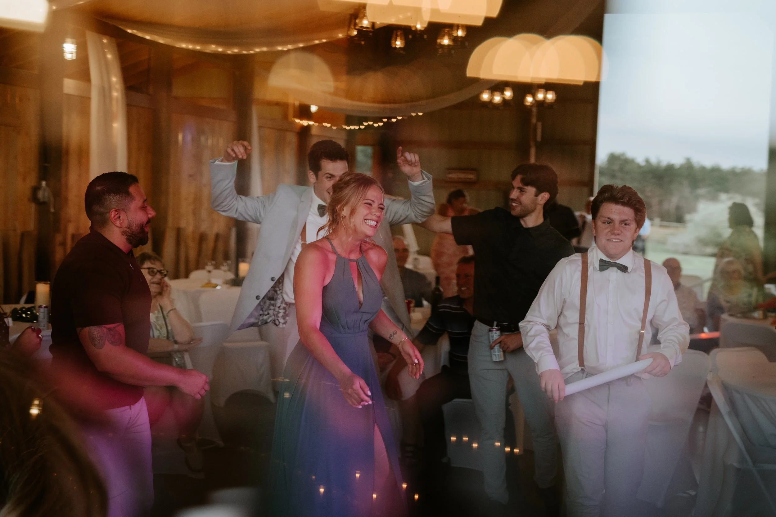 People celebrating at a wedding reception arn with string lights, with a woman dancing and smiling in a purple dress, surrounded by men, one in a light suit, another in black shirt, and a young man with suspenders holding a rolled paper, with guests 