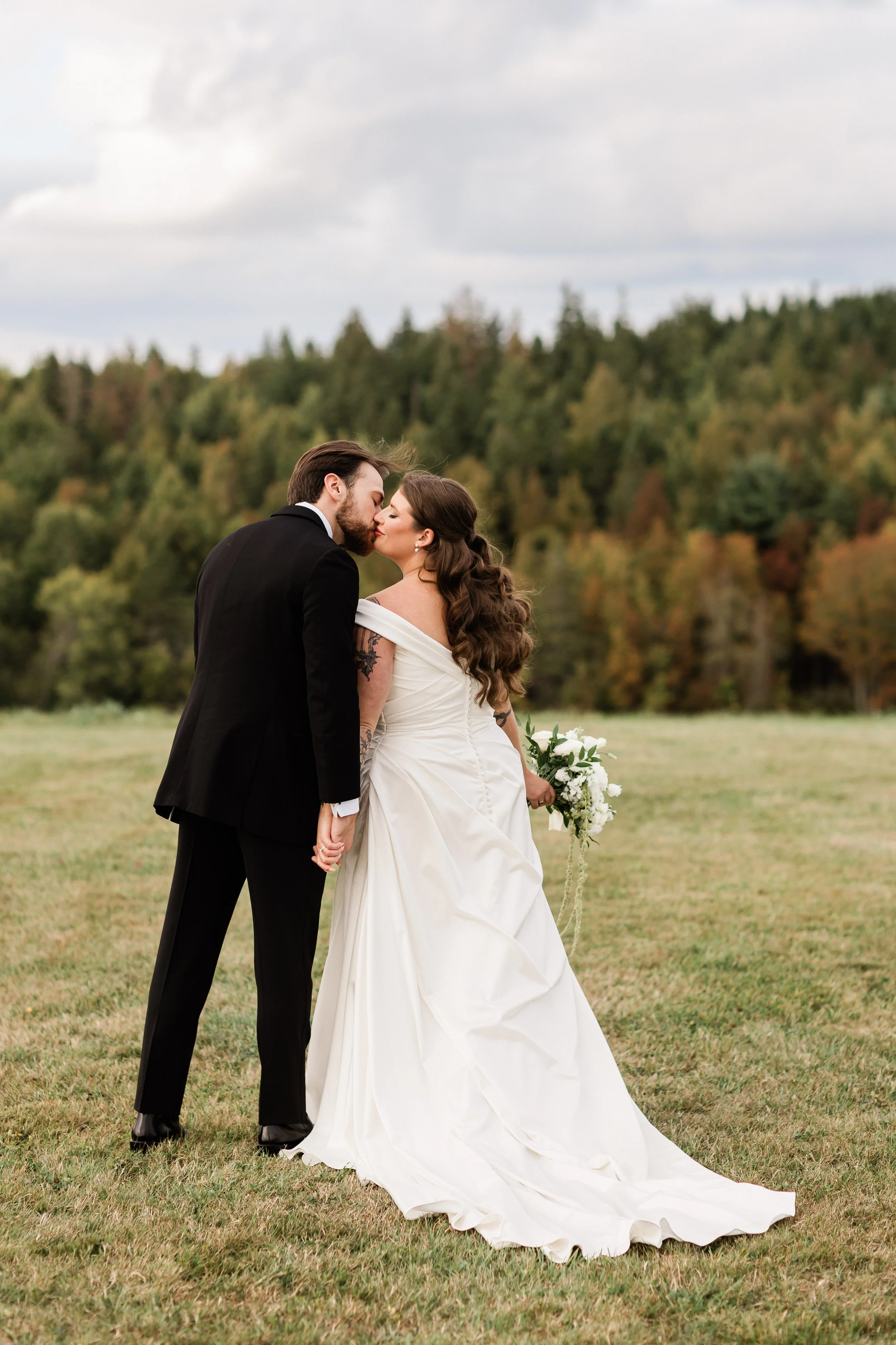 A bride and groom share a kiss outdoors on a grassy field, holding hands. The bride is dressed in a white wedding gown and holding a bouquet, while the groom is in a black suit. There is a forested area with trees in fall colors in the background.