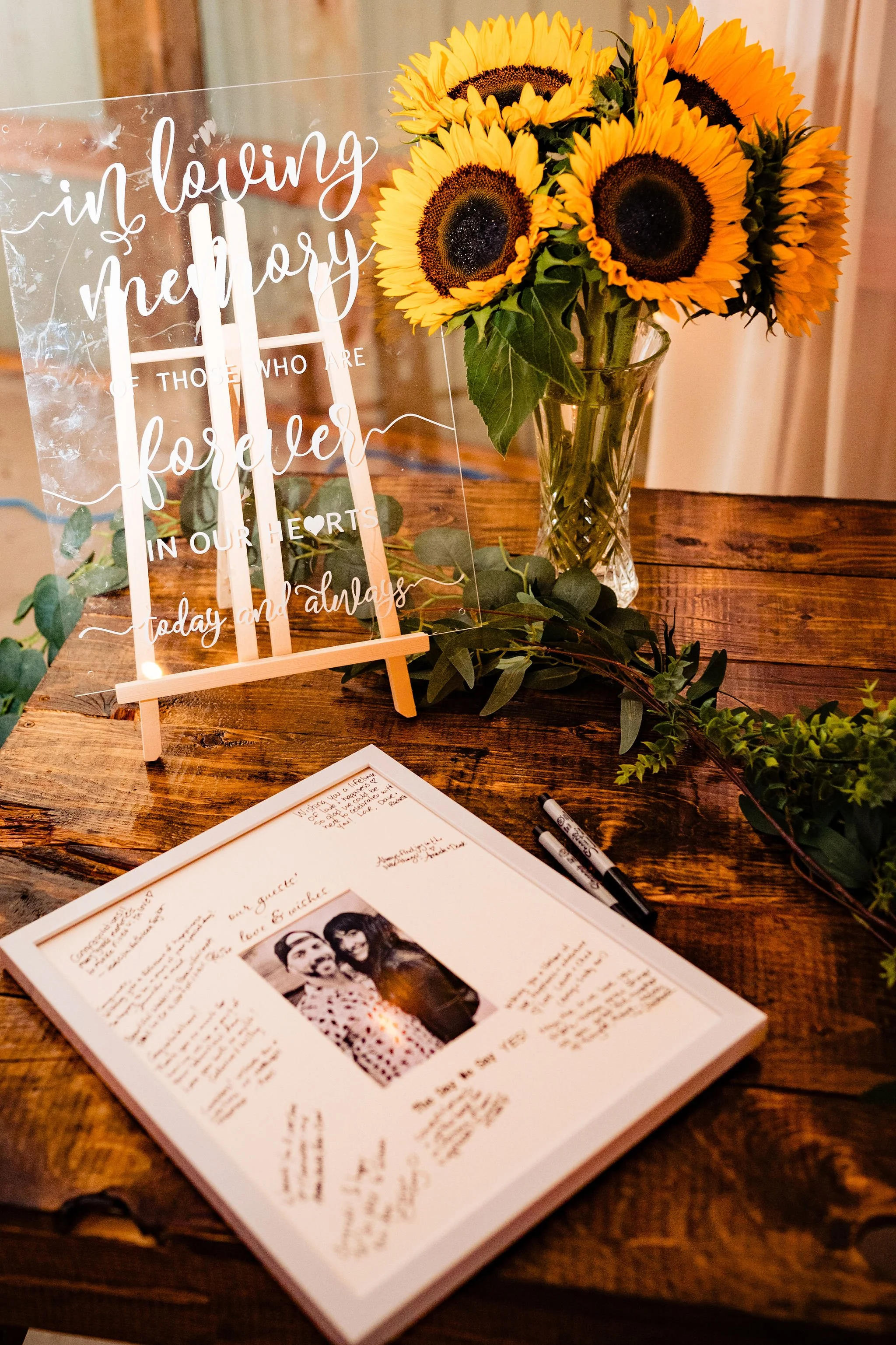 A wooden table with a framed handwritten note, a bouquet of sunflowers in a glass vase, a small easel with a transparent sign that reads 'In loving memory of those who are forever in our hearts, today and always,' and some pens and greenery.