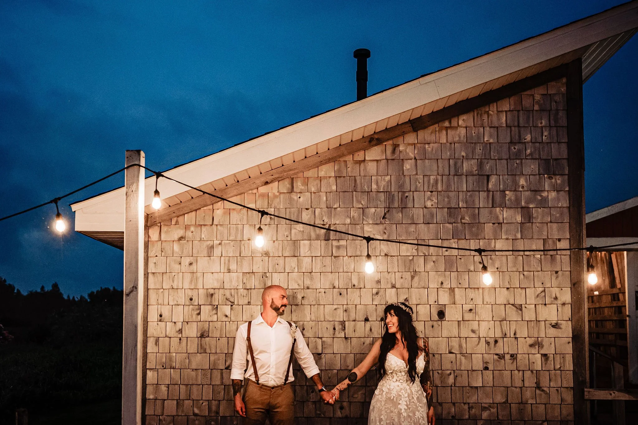 A couple holding hands, standing outdoors at night, with a wooden building and string lights in the background.