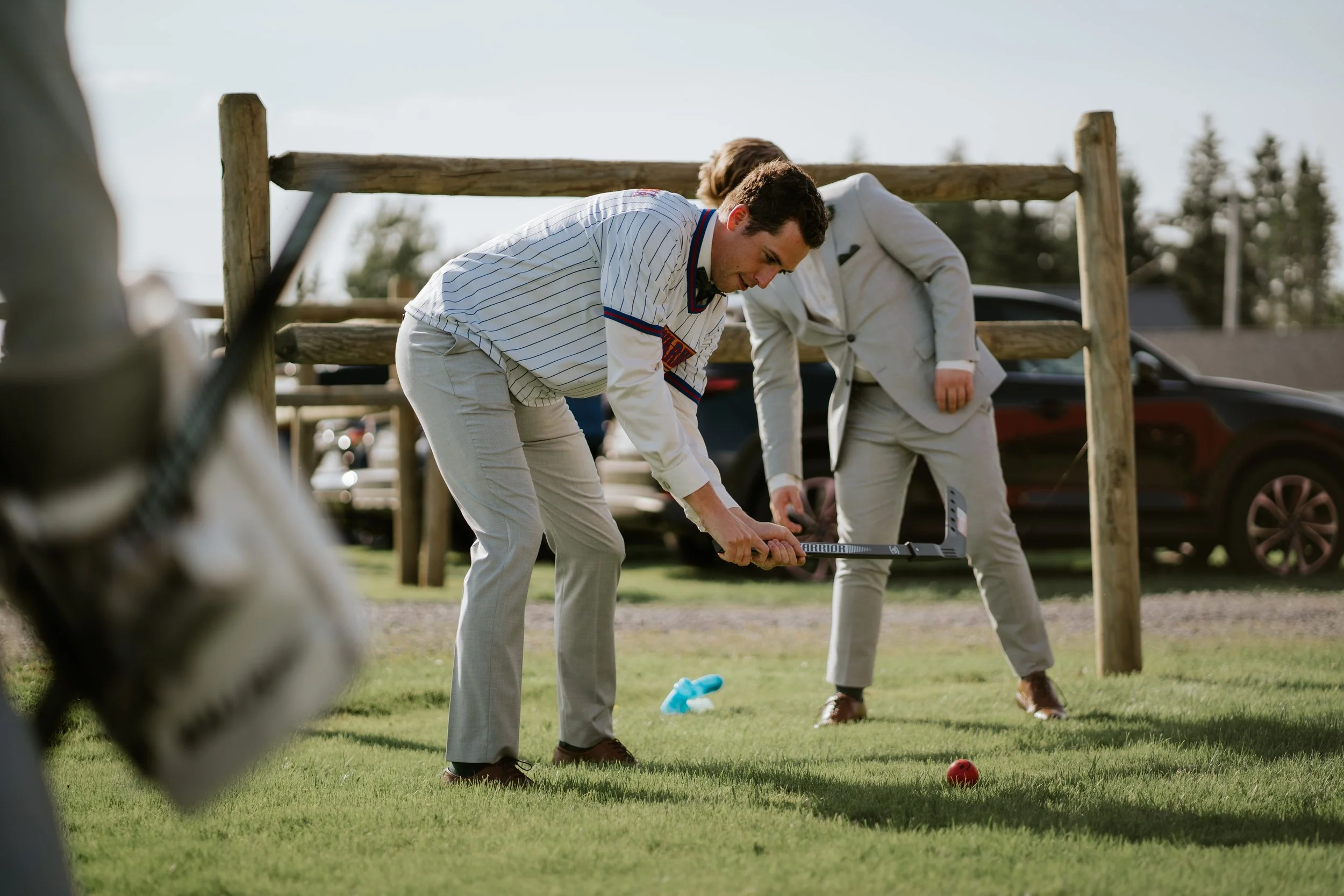 Two men in suits are playing hockey outdoors on a grassy field, one using a hockey stick to hit an orange ball.