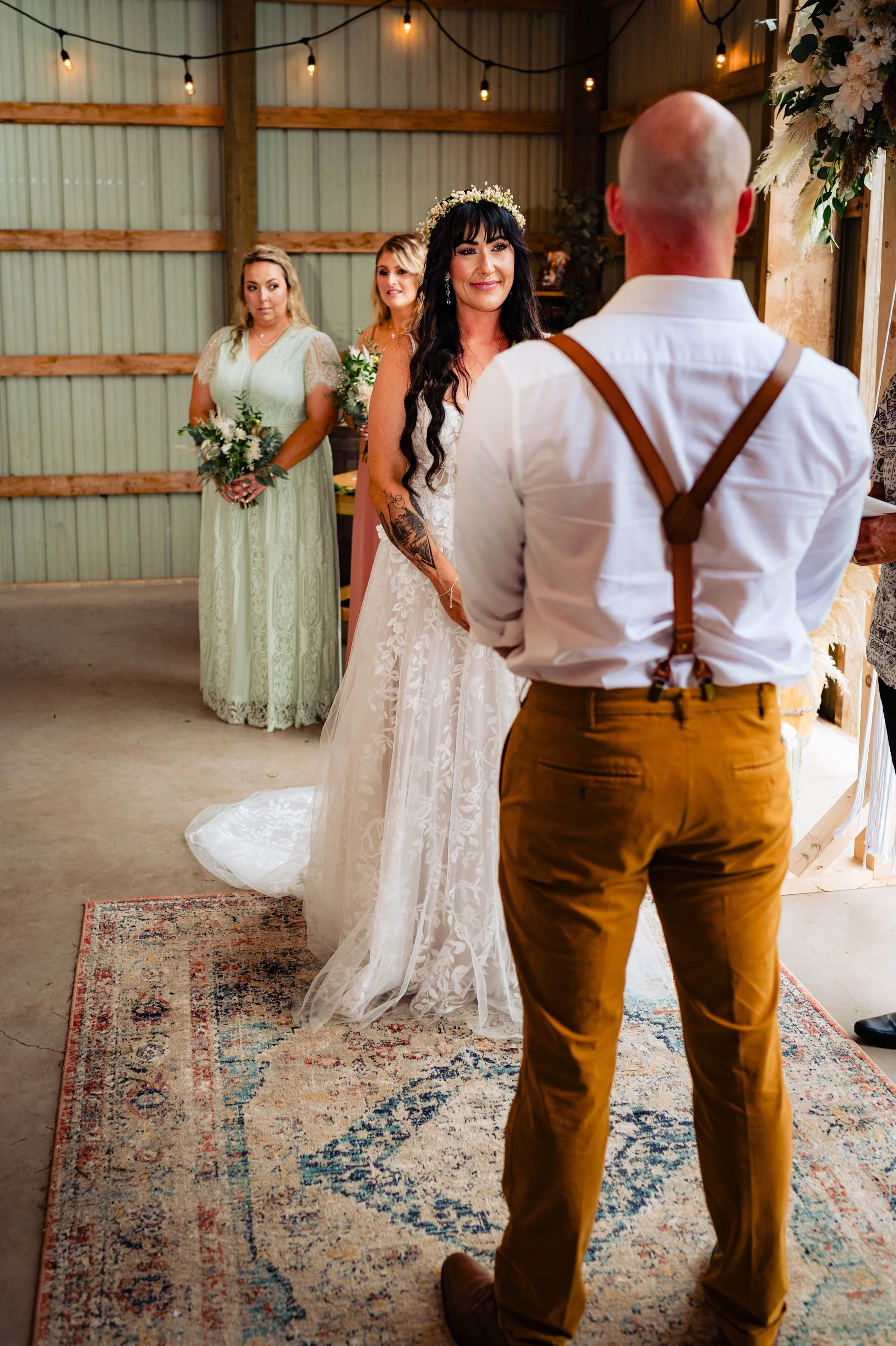 A wedding ceremony with a bride, groom, and two bridesmaids inside a rustic barn. The bride is wearing a white lace wedding dress with dark hair and a flower crown. The groom is facing away wearing a white shirt with suspenders. The bridesmaids are h