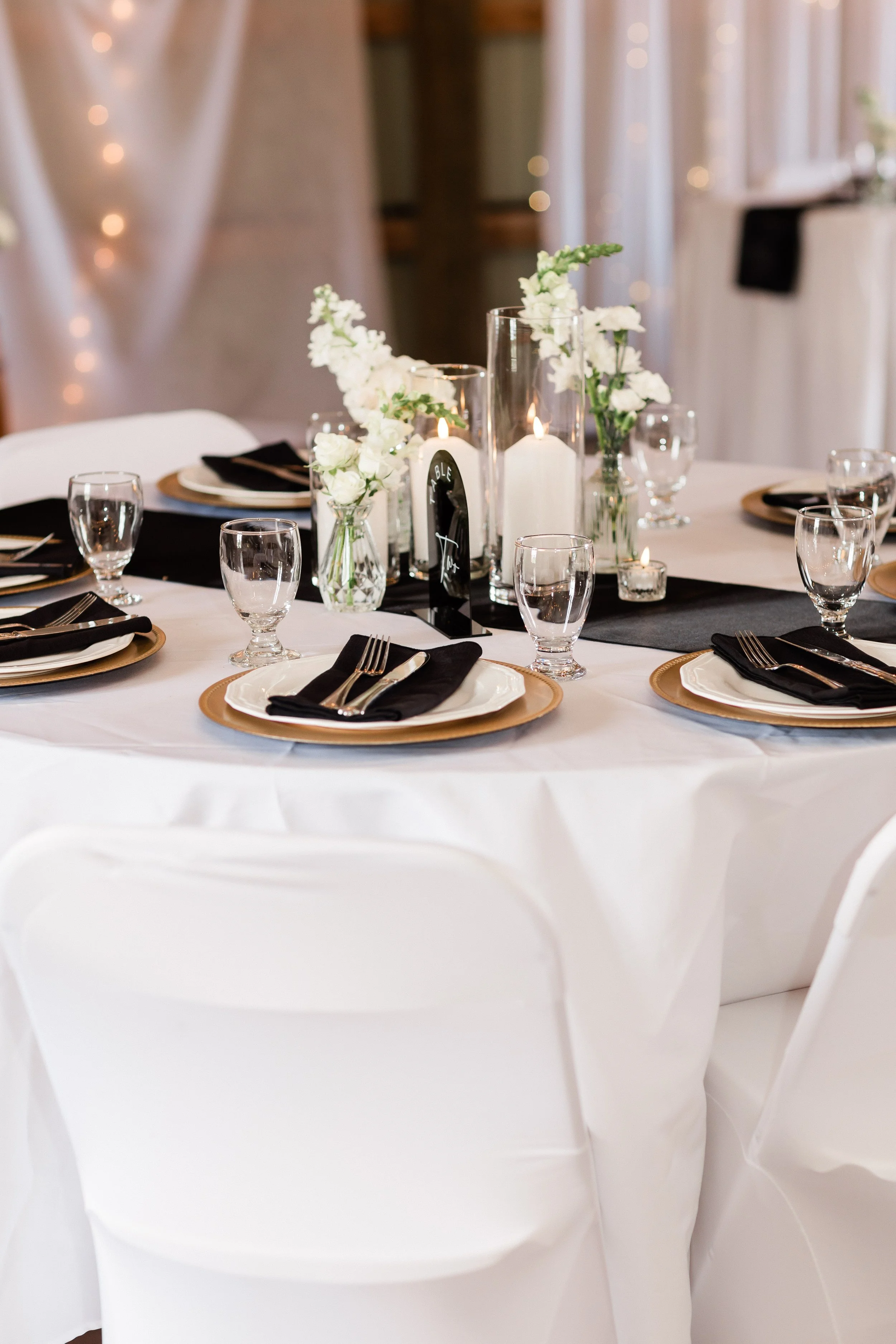 Elegant dinner table setting with white tablecloth, black napkins, gold-rimmed plates, clear water glasses, and floral centerpieces with white flowers, candles, and a black table runner.
