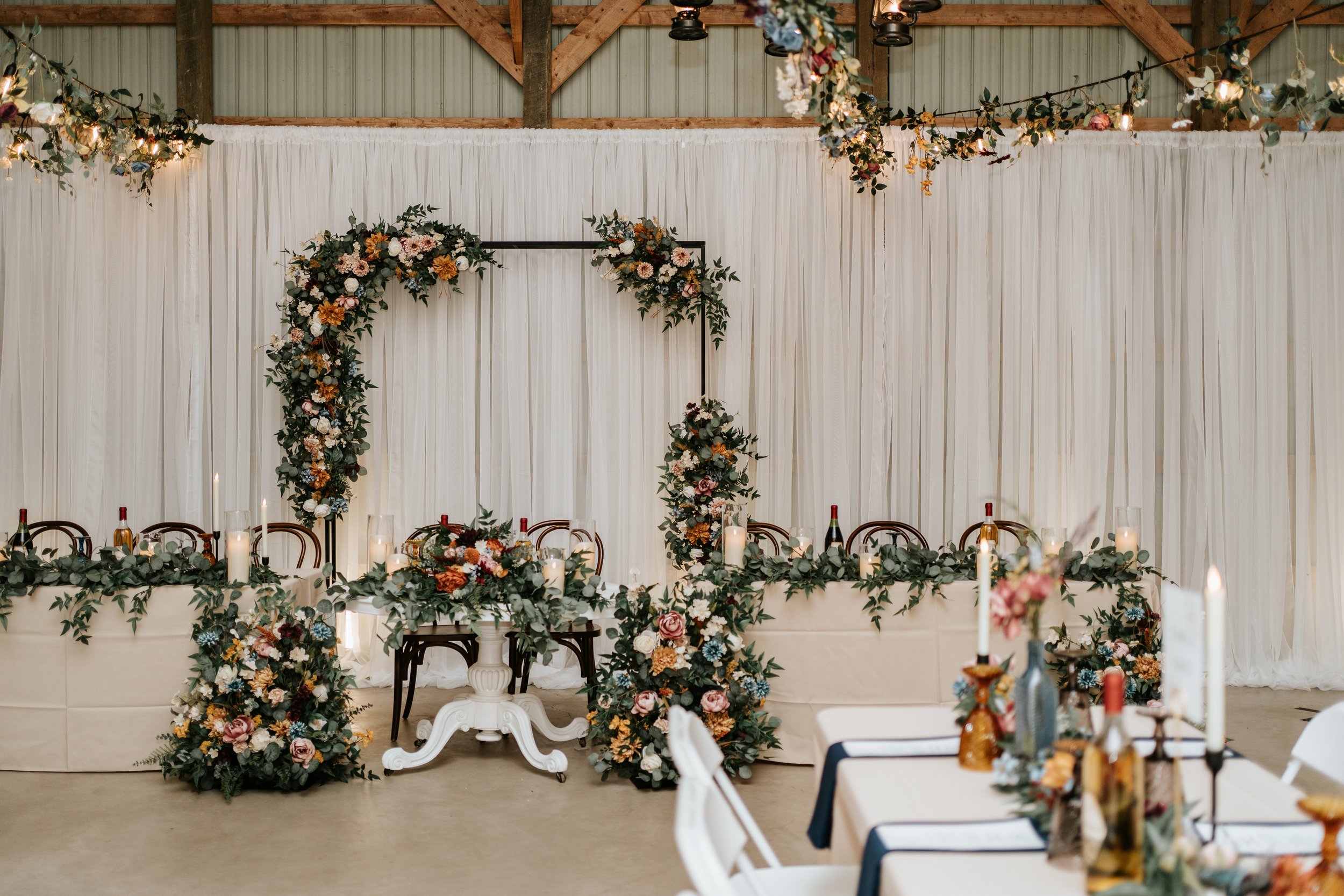 Wedding reception setup with floral arch, decorated table, candles, and bottles in rustic venue.