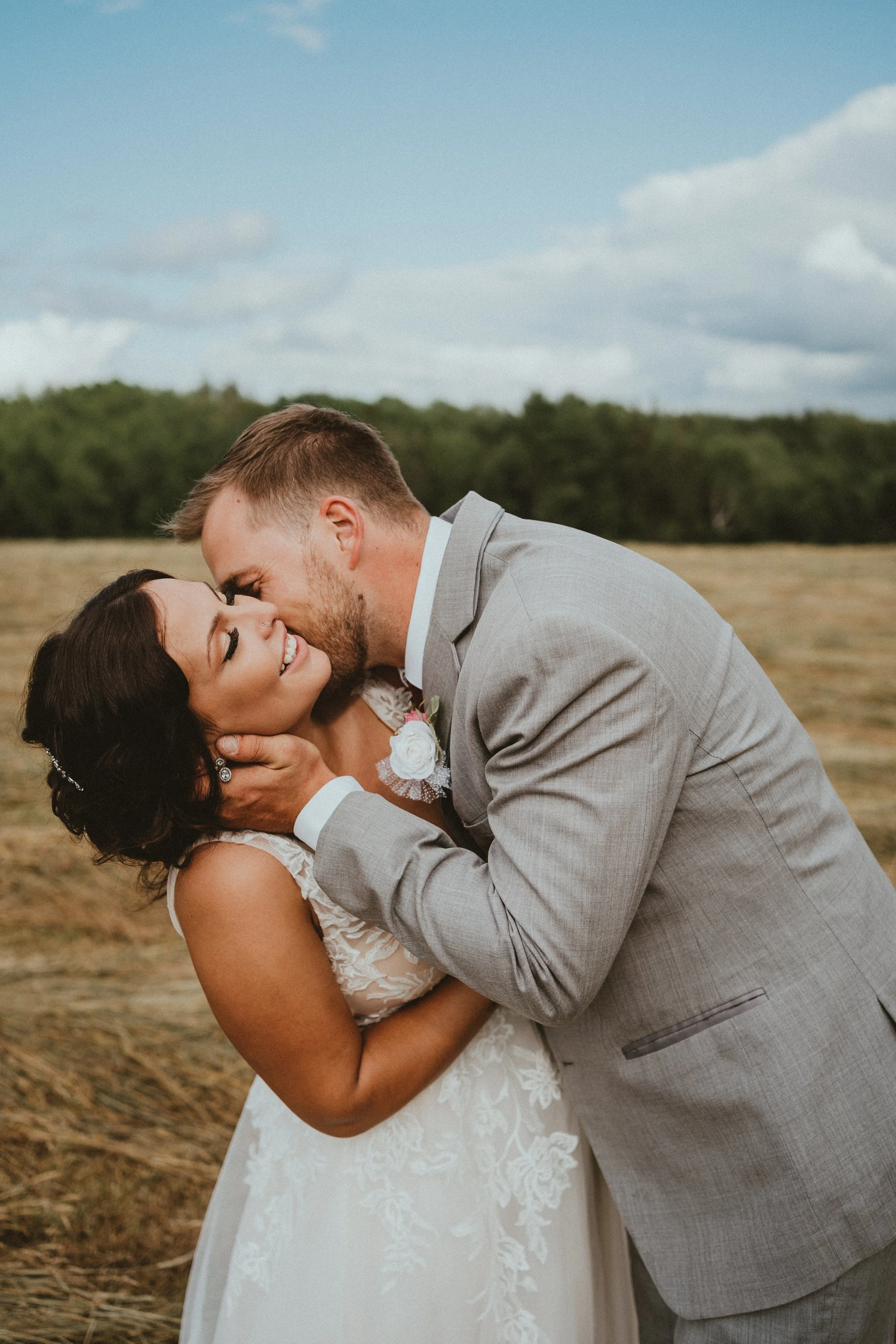 A bride and groom sharing a romantic kiss outdoors on their wedding day, with open field and trees in the background.