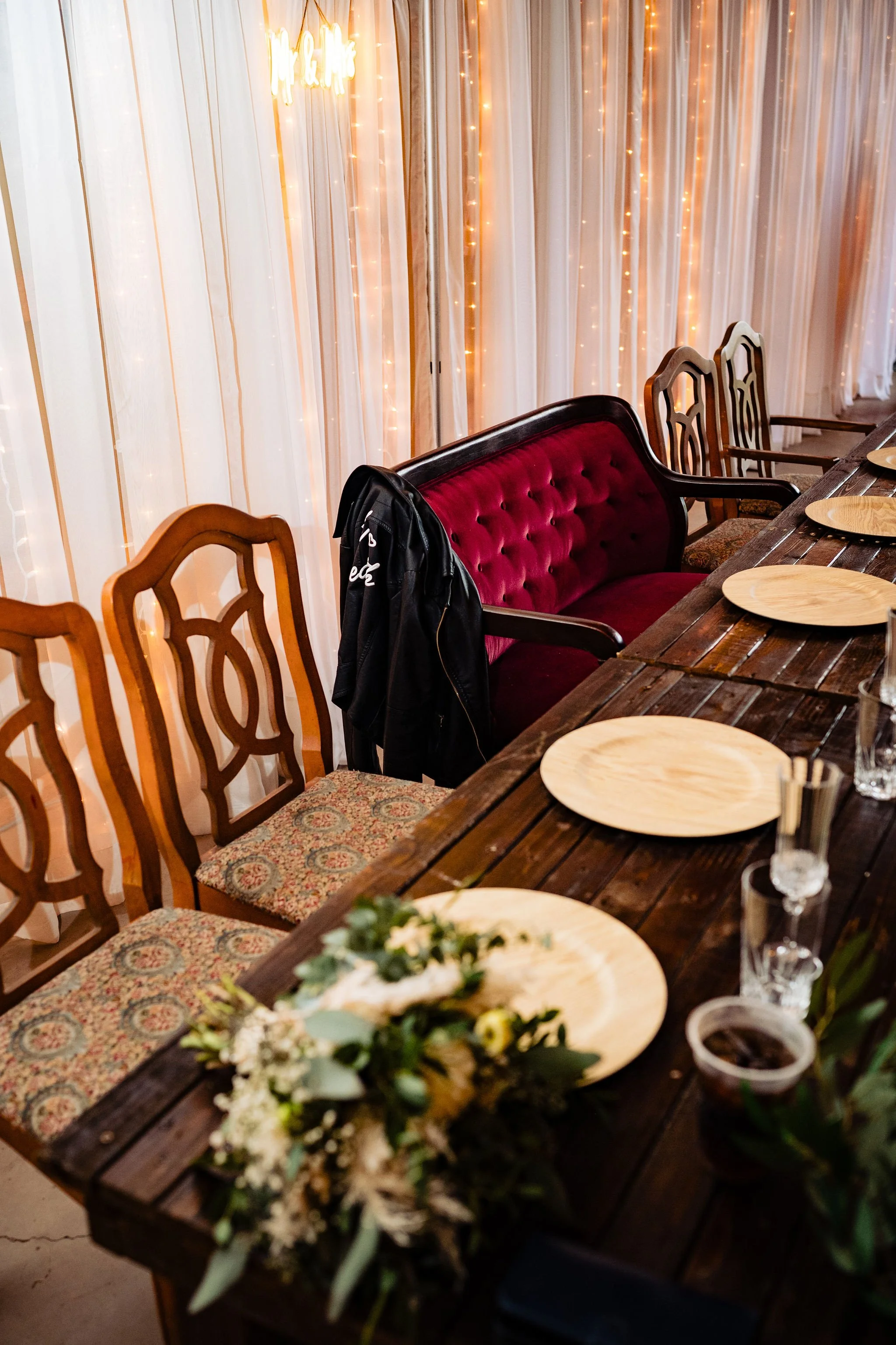 A decorated dining table with wooden chairs, a maroon velvet couch, plates, glasses, and a floral centerpiece, set against a backdrop of string lights and curtains for a celebration or special event.