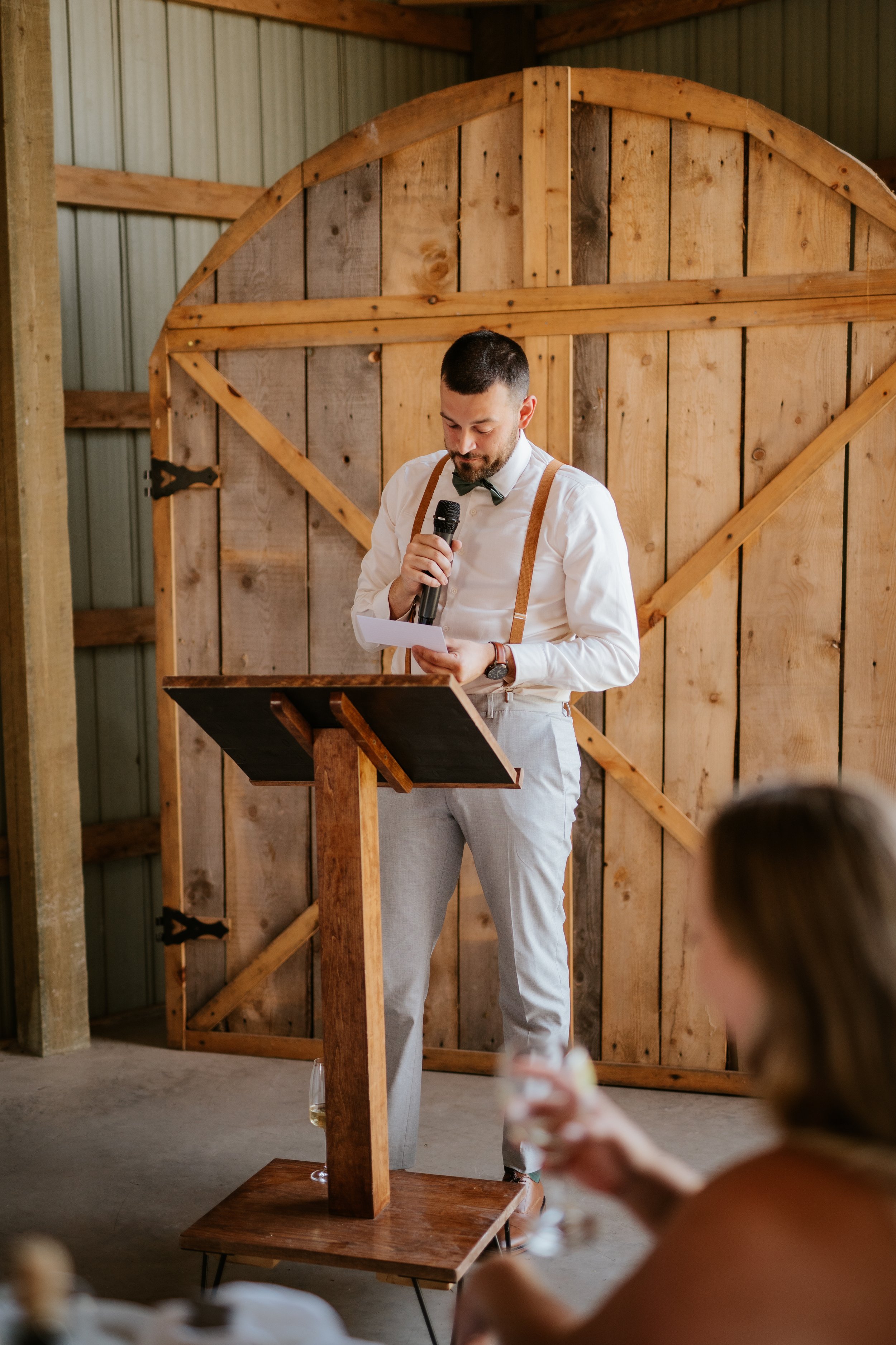 A man giving a speech at a wedding reception, standing at a wooden podium with a microphone, wearing a white shirt, suspenders, and light gray pants, at Parker's Barn.