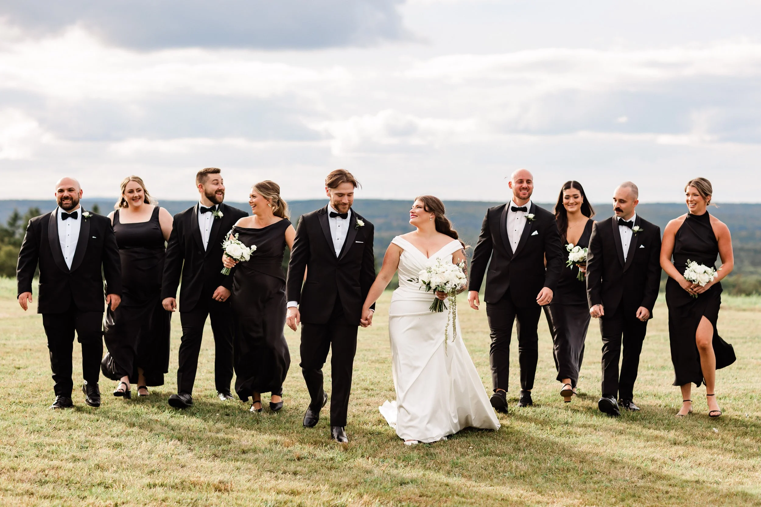 A wedding party of eleven people walking across a grassy field, with the bride and groom in the center, holding hands and smiling, surrounded by groomsmen and bridesmaids dressed in black and wearing black shoes, with a countryside landscape and clou