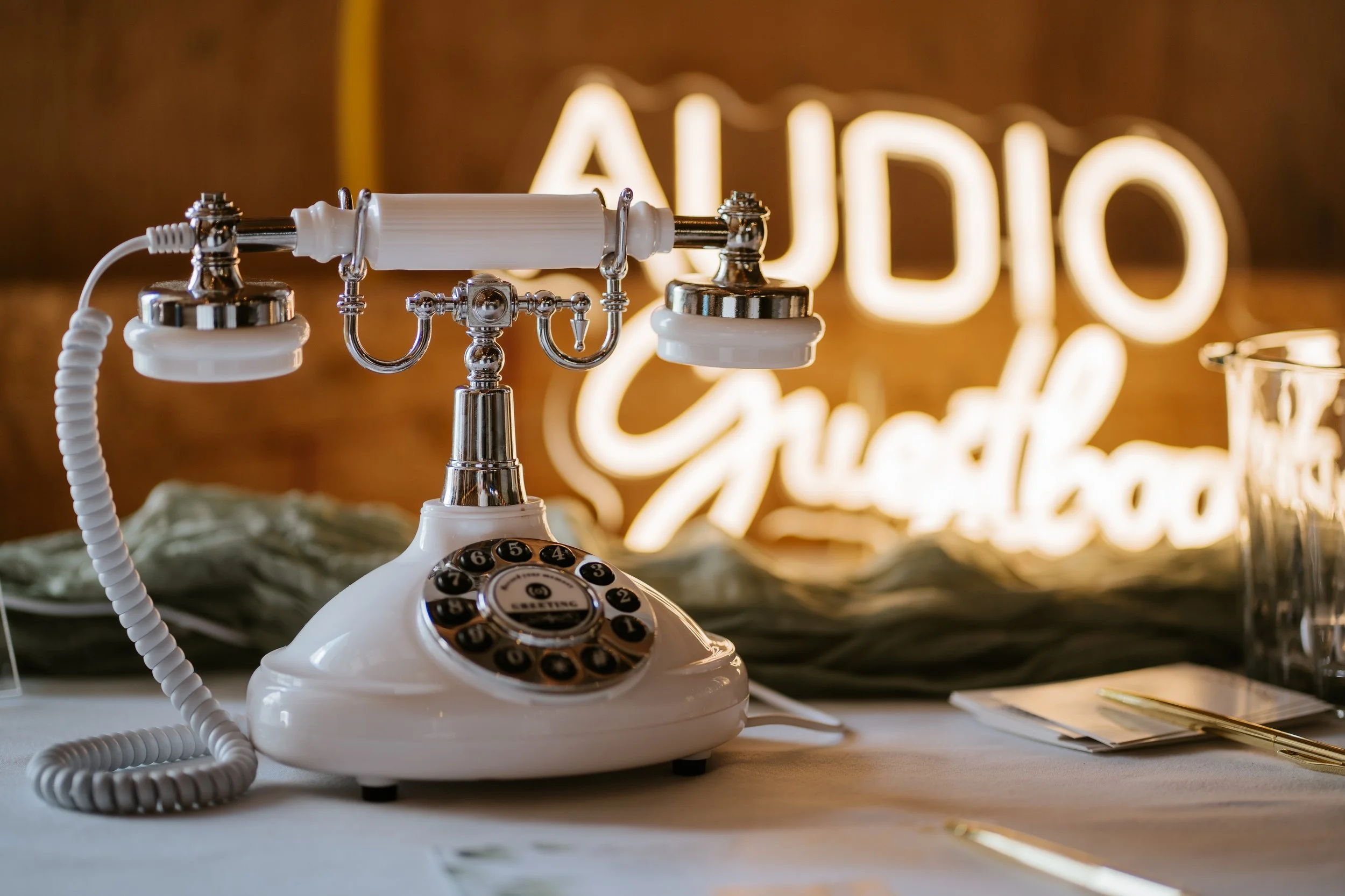 Vintage white rotary telephone on a table with a blurred background featuring a neon sign that says 'AUDIO CRAFT'.