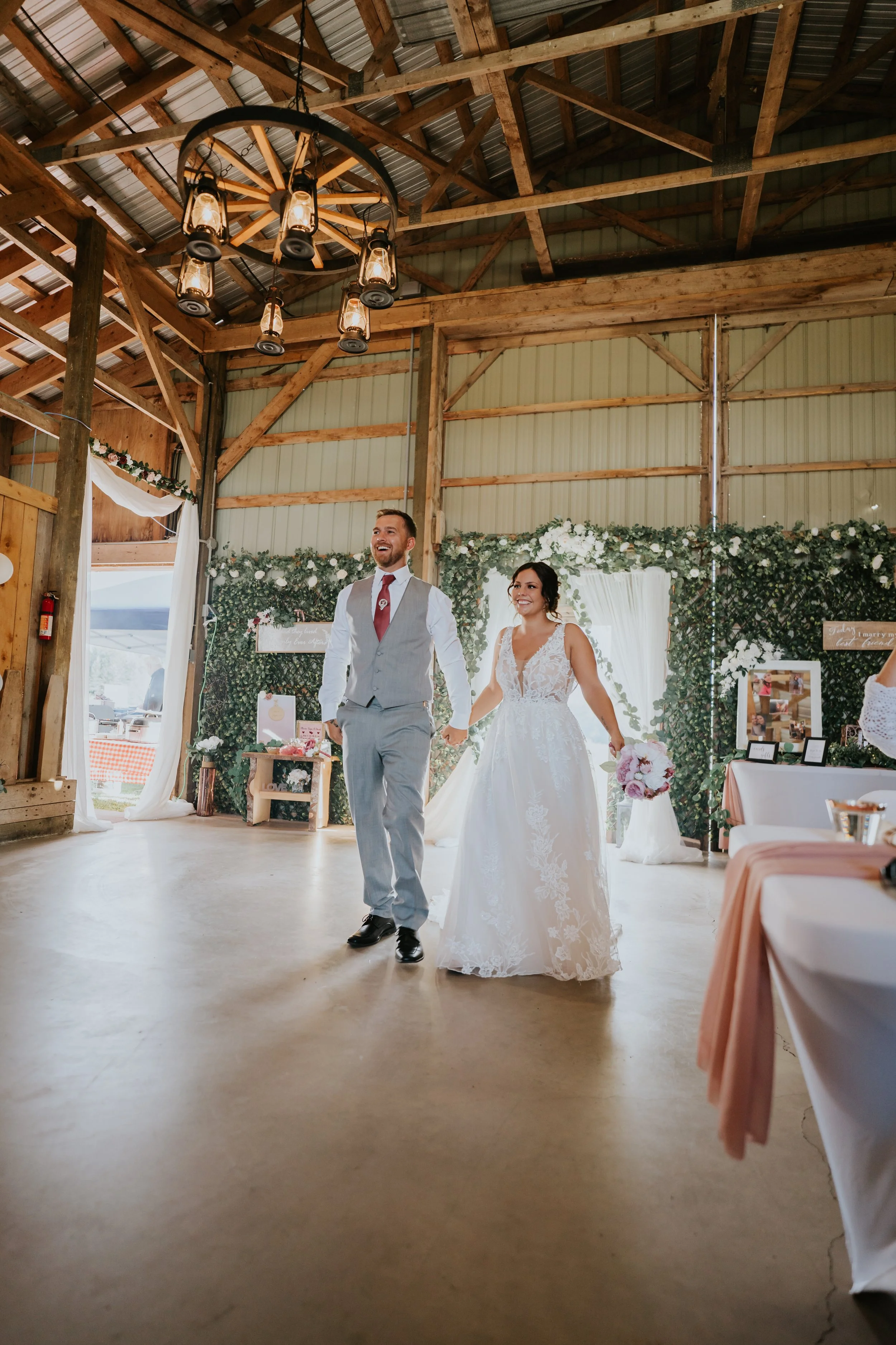 A newlywed couple walking hand in hand into their wedding reception in a rustic barn. The bride is in a white lace wedding gown holding a bouquet, and the groom is in a gray vest and pants with a white shirt and red tie. The barn features wooden beam