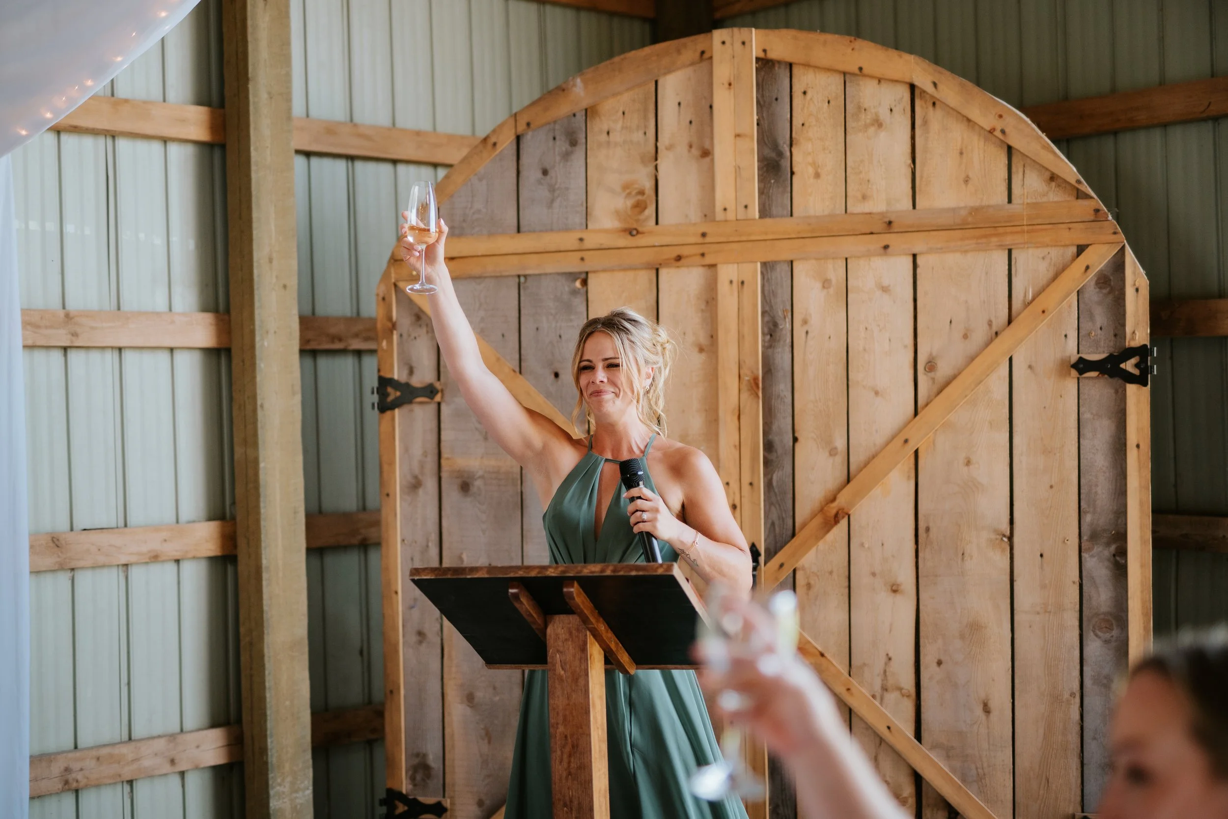A woman in a green dress giving a toast with a glass of champagne at a wooden wedding reception.