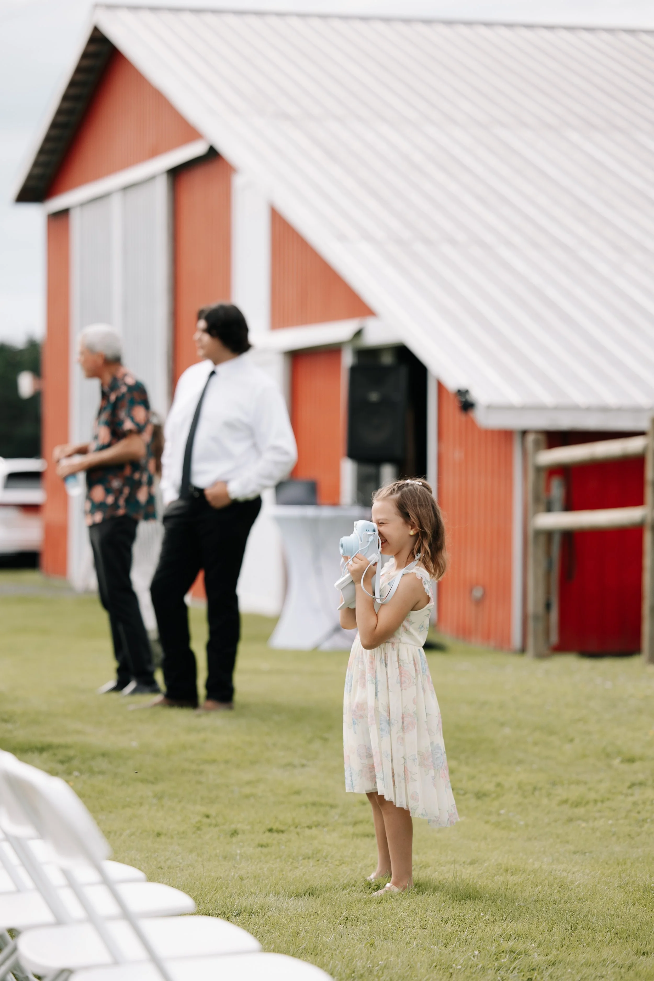 A young girl in a floral dress holding a camera to her eye, smiling, at an outdoor event with a red barn in the background, and two men in formal attire standing nearby.