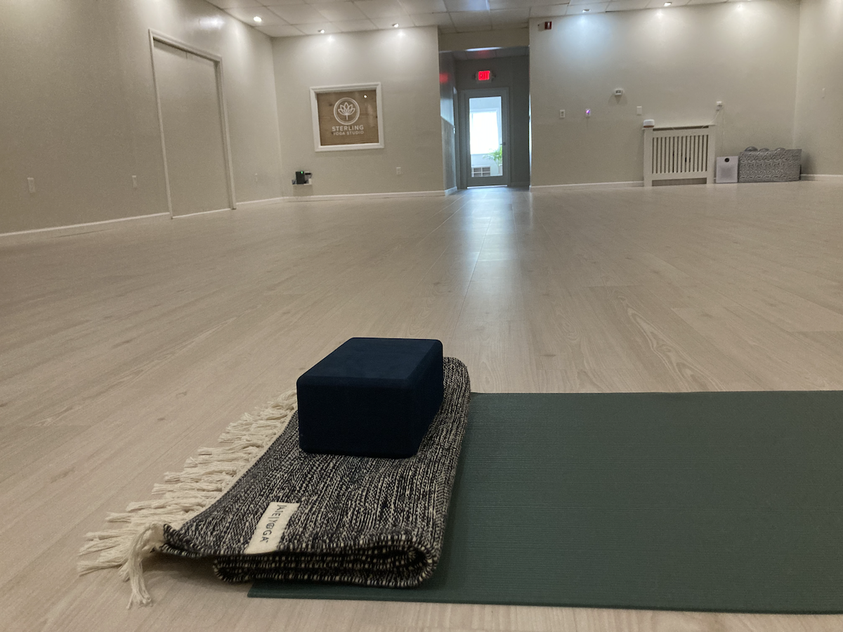 Empty yoga studio with a yoga mat, a block, and a blanket in the foreground, and a door leading outside in the background.