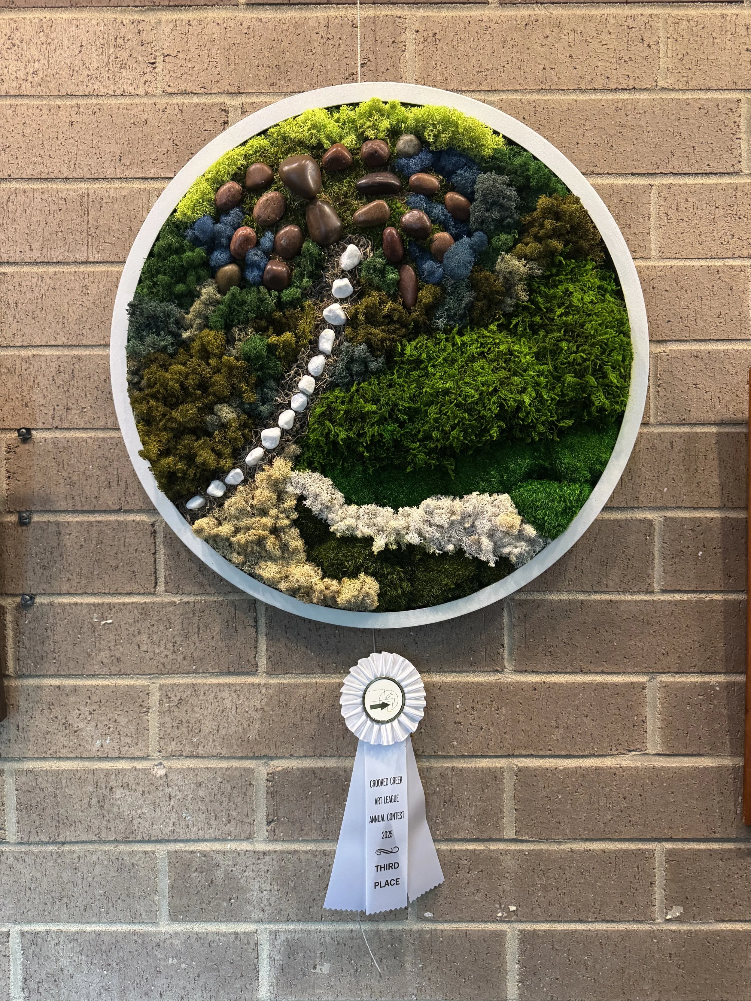 A circular artwork made of various mosses, rocks, and plants, with a path of white stones, mounted on a brick wall. A ribbon at the bottom indicates this is a third-place award in a 2023 art contest.