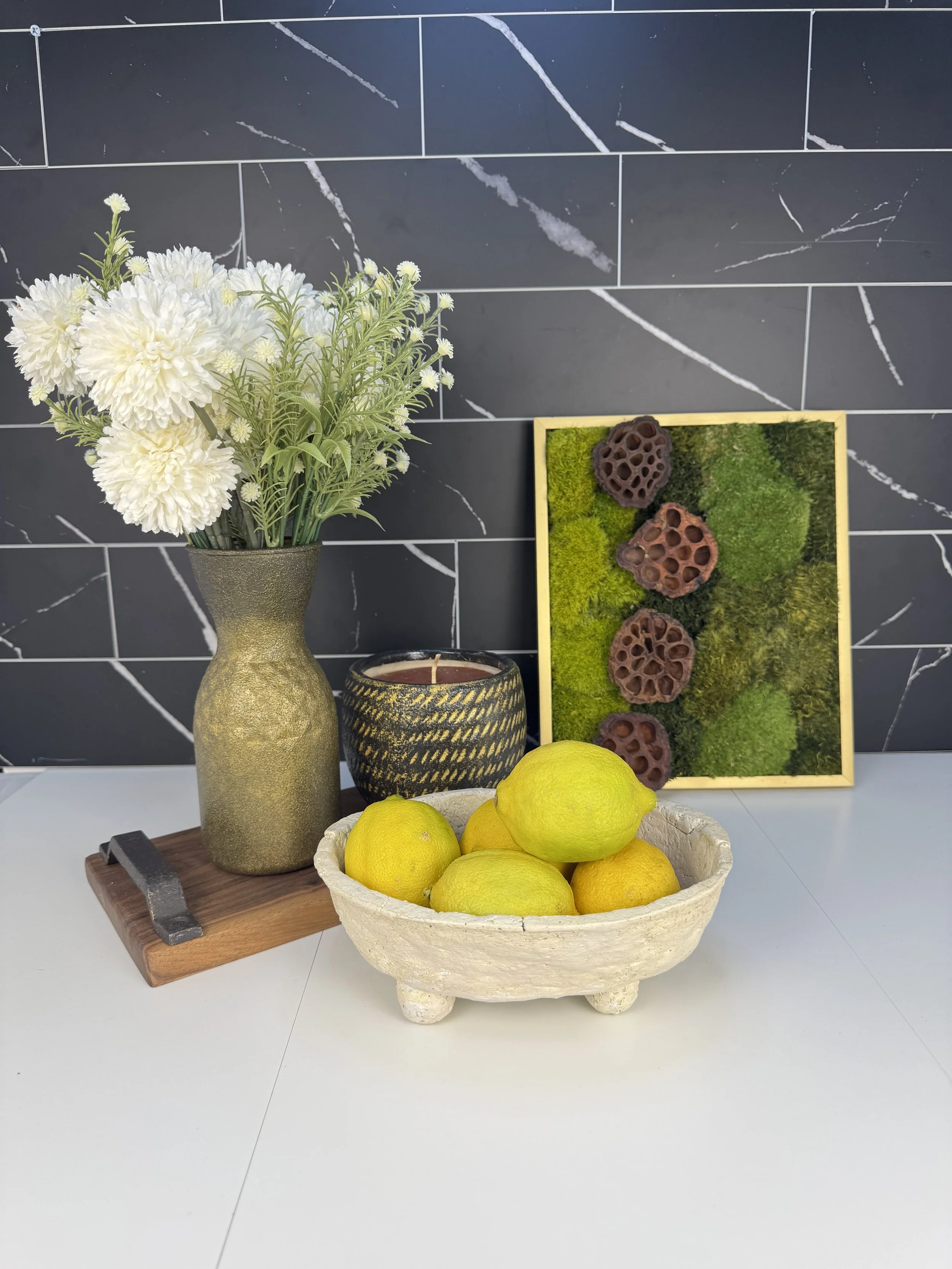A white bowl with lemons and limes on a white surface, a black and gold decorative candle, a tall gold vase with white flowers, a framed piece of green moss and succulents, and a wooden cutting board with a small black handle, all set against a black marble tiled wall.