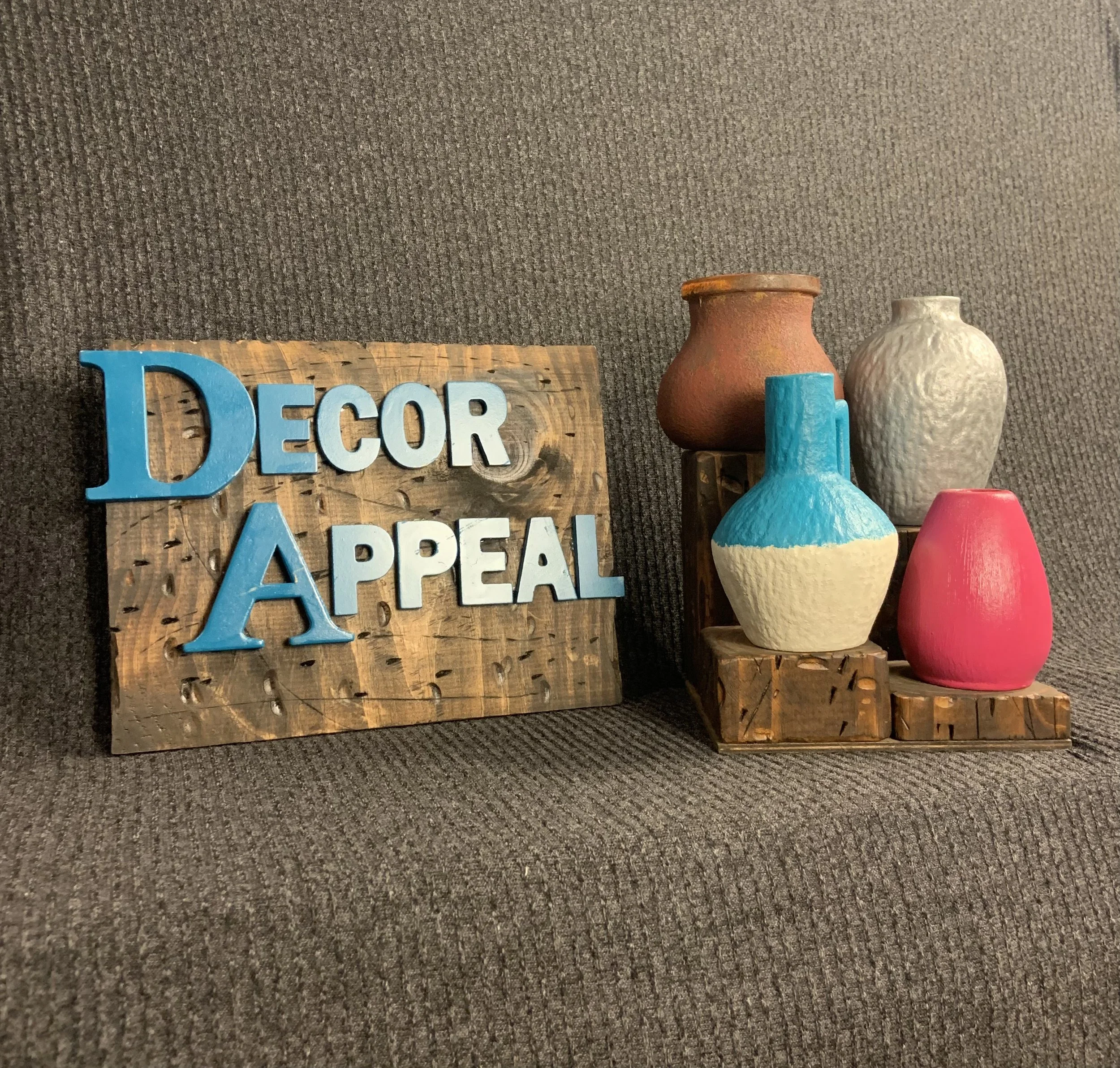 Decorative sign reading 'DECOR APPEAL' next to a display of four colorful vases on wooden stands against a brown textured background.