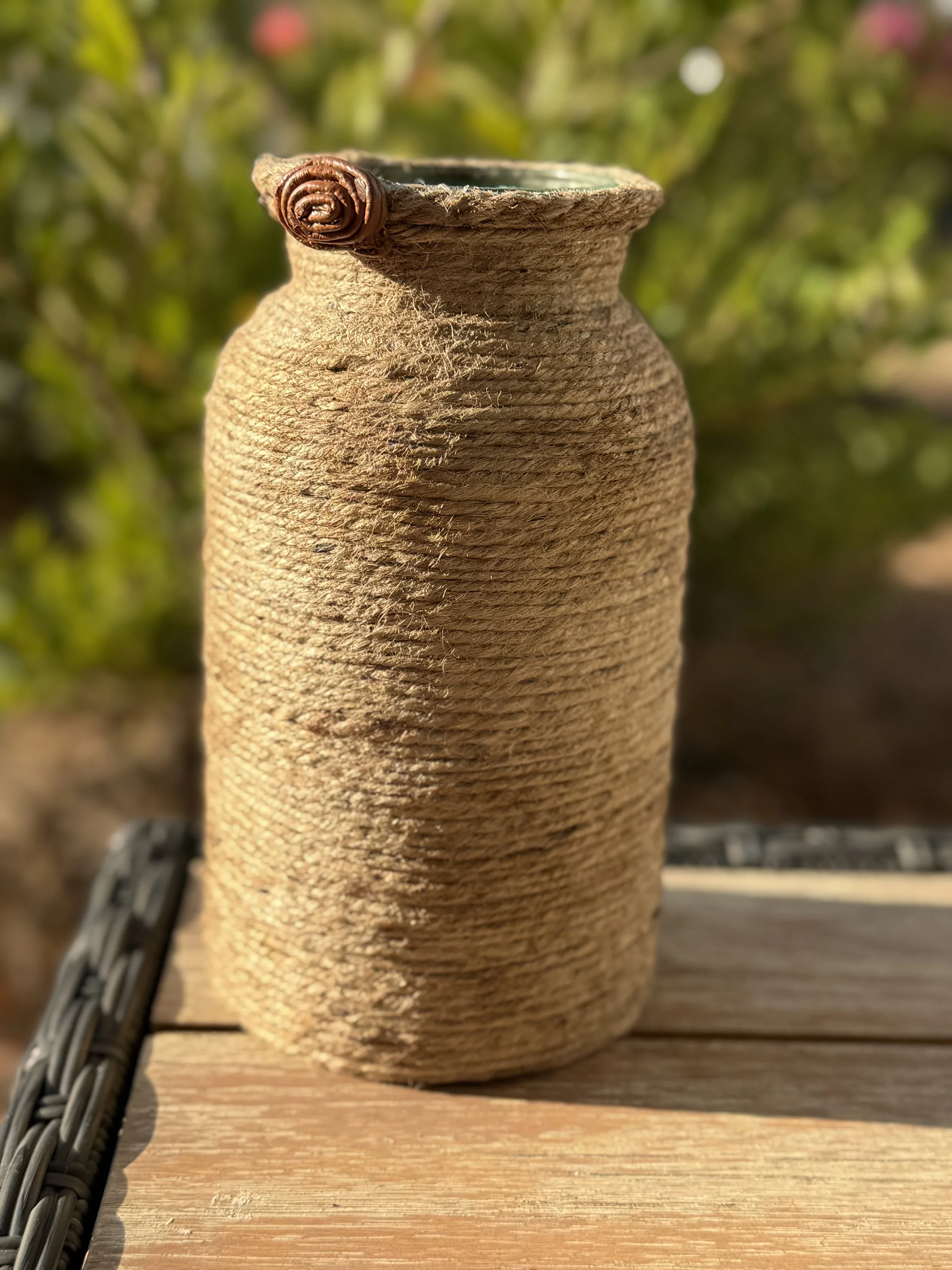 A beige ceramic vase with a textured surface, featuring a twisted clay decoration on the rim, placed on a wooden surface outdoors with blurred greenery in the background.