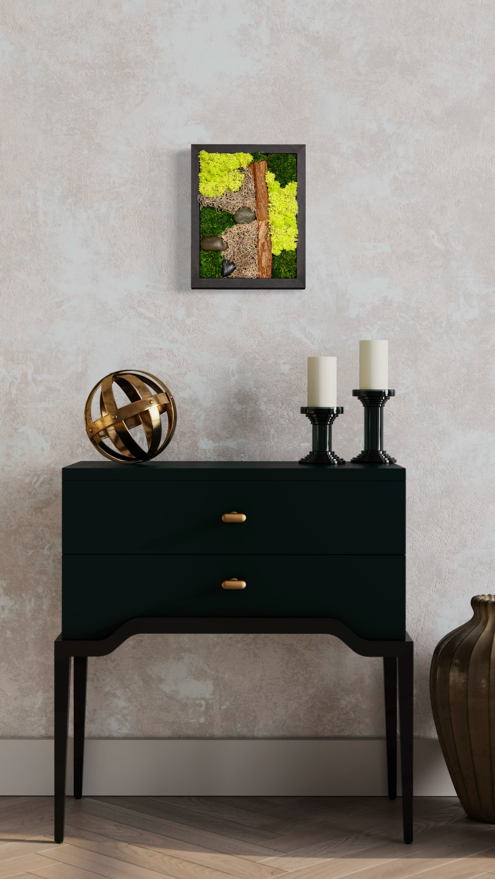 Wall art with moss, stones, and wood on a black frame, placed on a wall above a black table with decorative objects including a golden geometric sphere and two black candle holders with white candles.