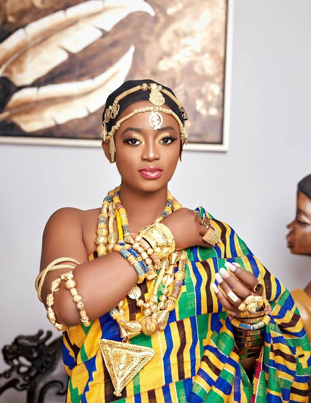 A woman dressed in traditional African attire with gold jewelry, including necklaces, rings, and headpieces. She is wearing a colorful kente cloth and is looking at the camera with a confident expression.