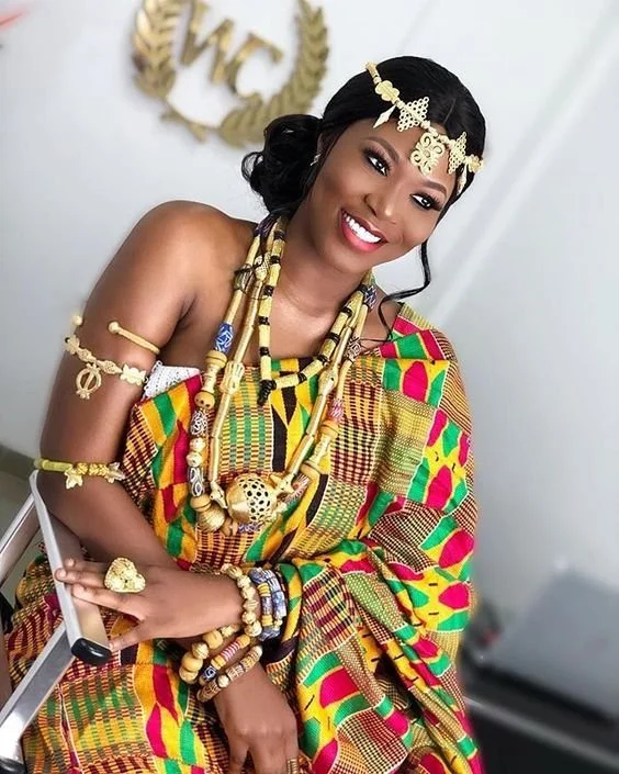 Woman dressed in colorful traditional African attire and jewelry, smiling, with a background featuring the letters 'WC' in gold.