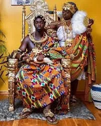 Two people dressed in traditional African attire seated on a decorative throne, with a yellow background and a patterned floor.