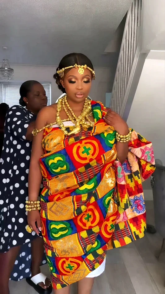 Woman dressed in traditional colorful Kente cloth with gold jewelry, standing indoors. Another woman in polka dot dress is assisting her.
