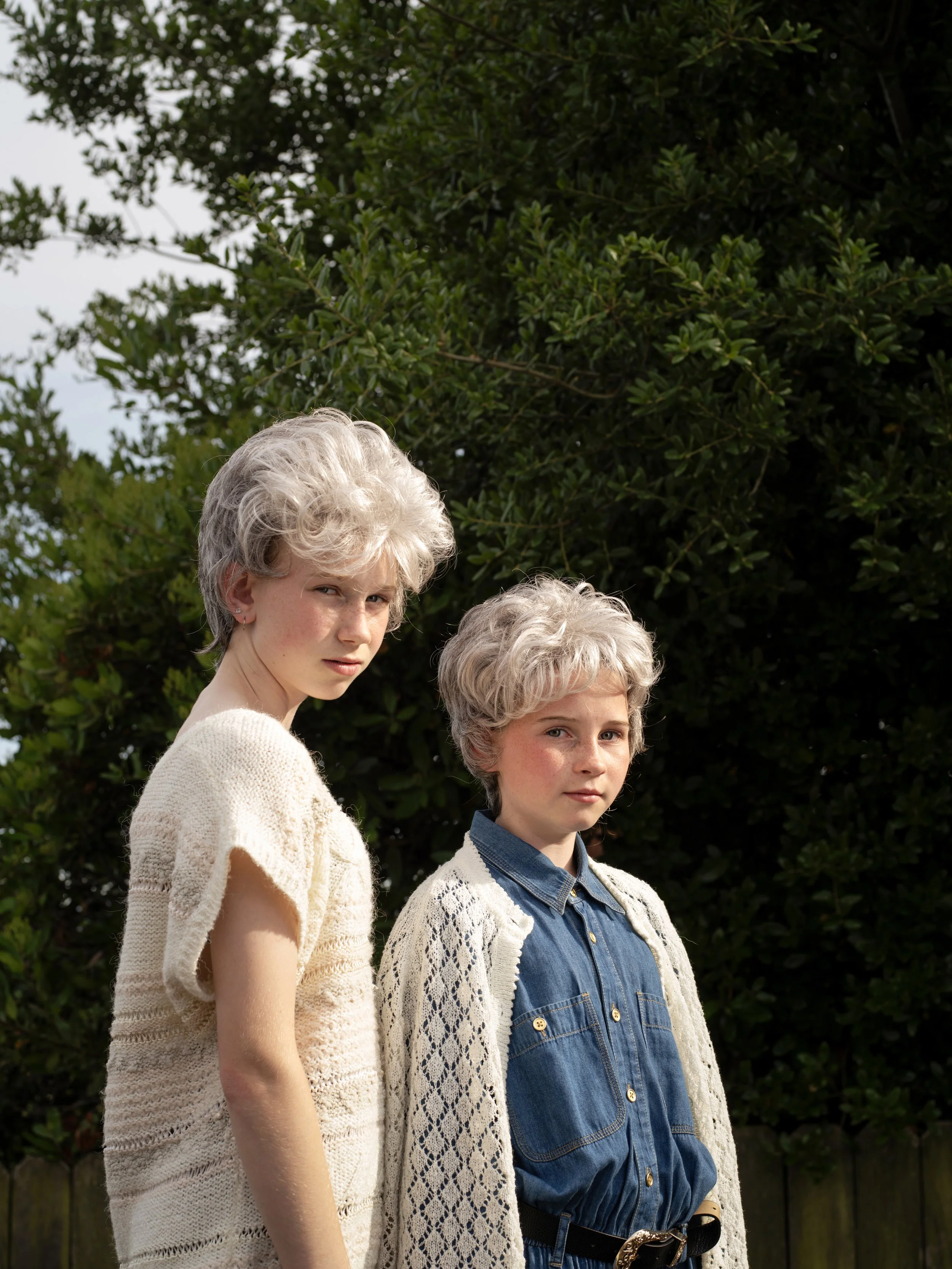 Two young women with gray curly hair standing outdoors in front of greenery, wearing vintage-style clothing, looking at the camera with serious expressions.
