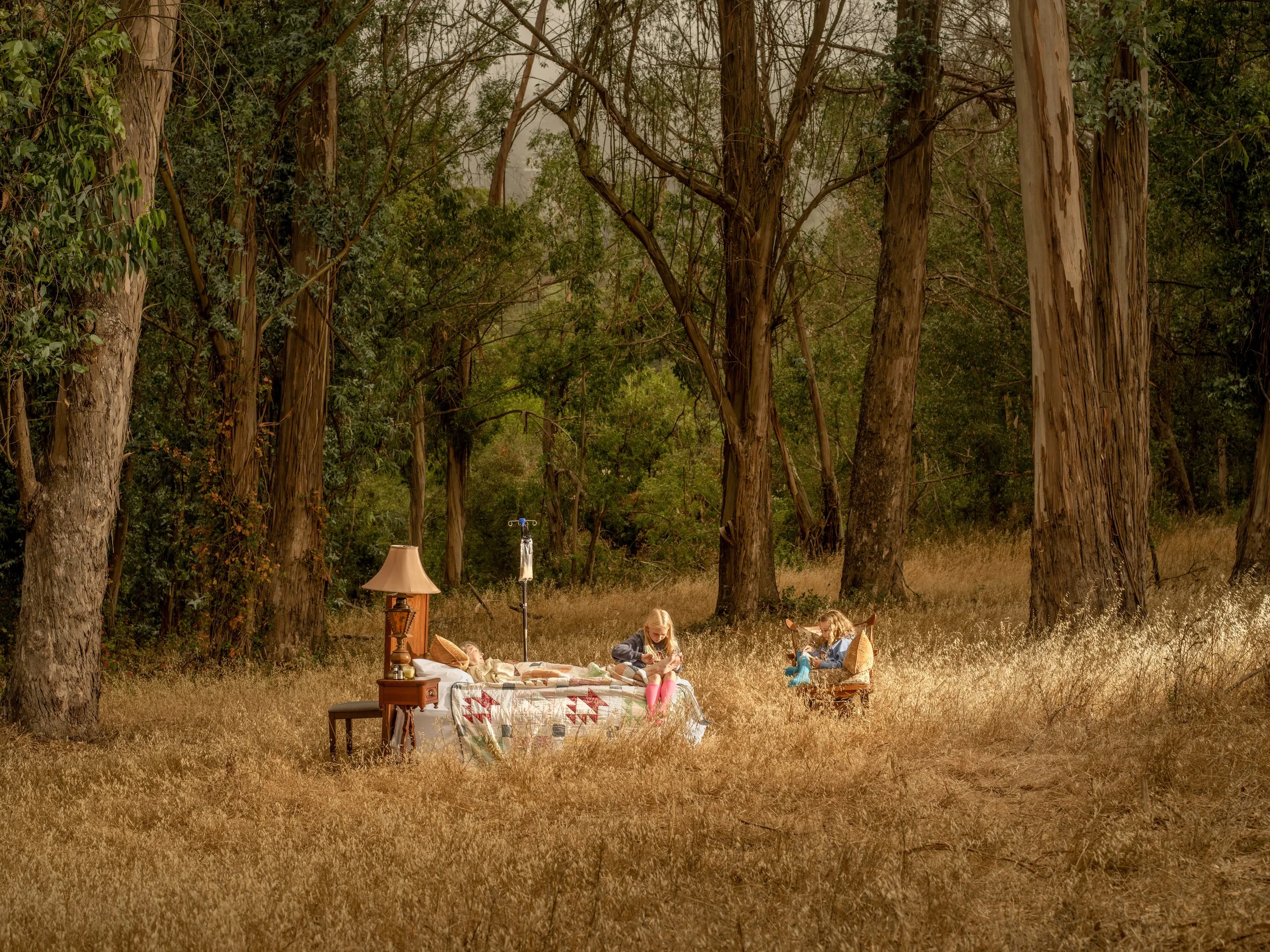 Children sitting outdoors in a forest, surrounded by tall trees and grass, with a table, lamp, and IV stand nearby.