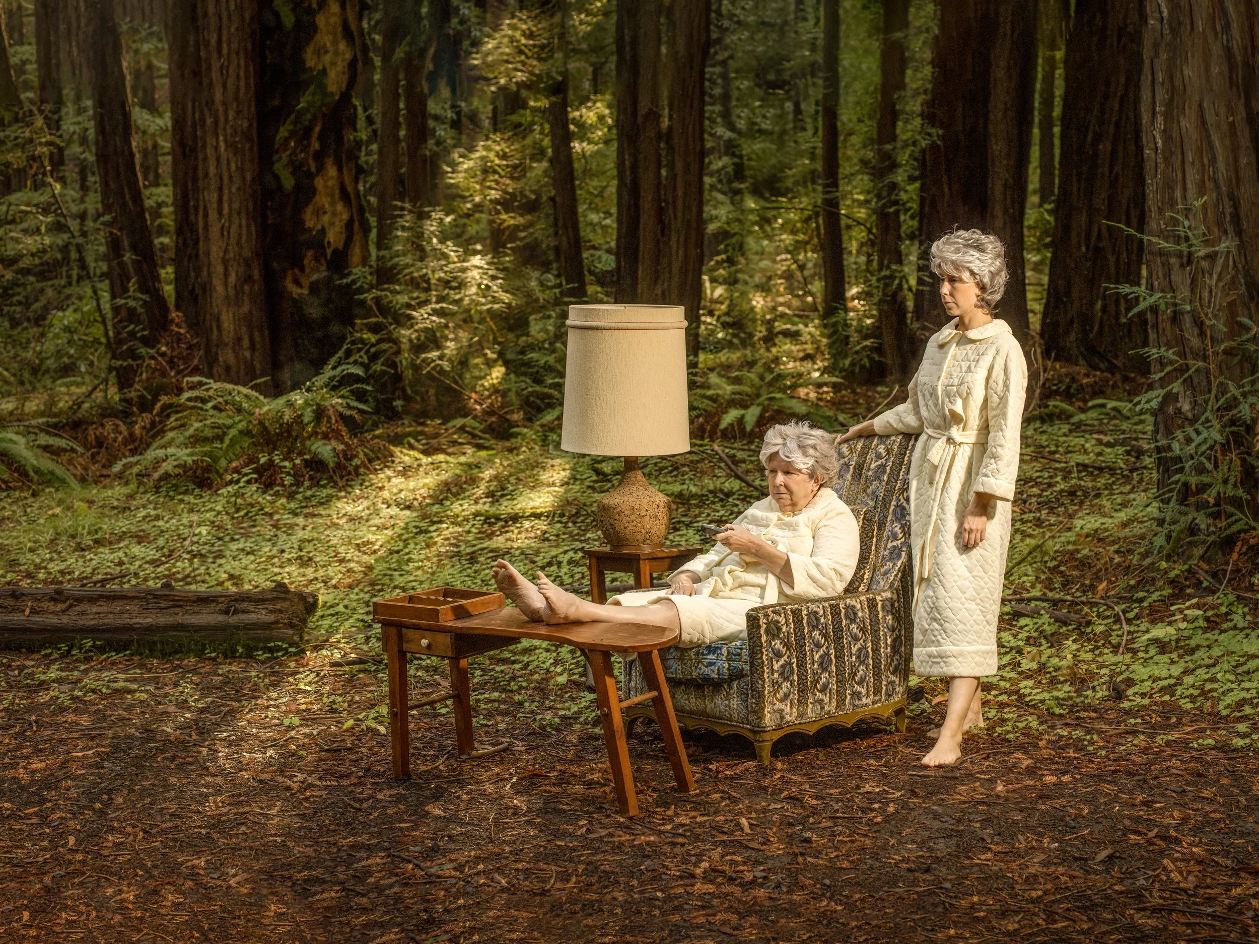 Two older women with gray hair in pajamas sitting and standing on a vintage armchair in a forest, with a small wooden table and a lamp nearby.