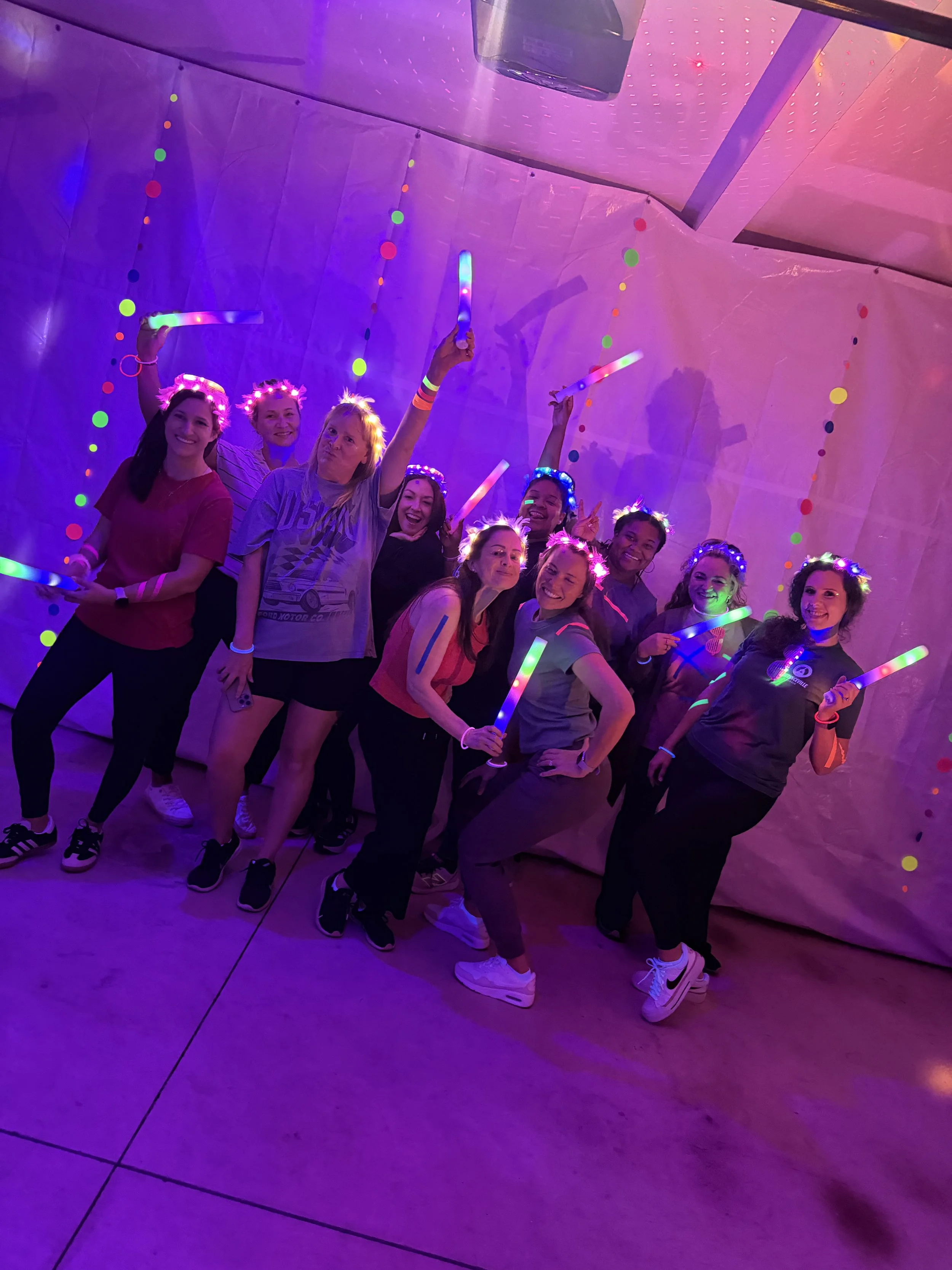Group of people at a glow-in-the-dark party holding glowing sticks, illuminated by purple, pink, and green lighting.