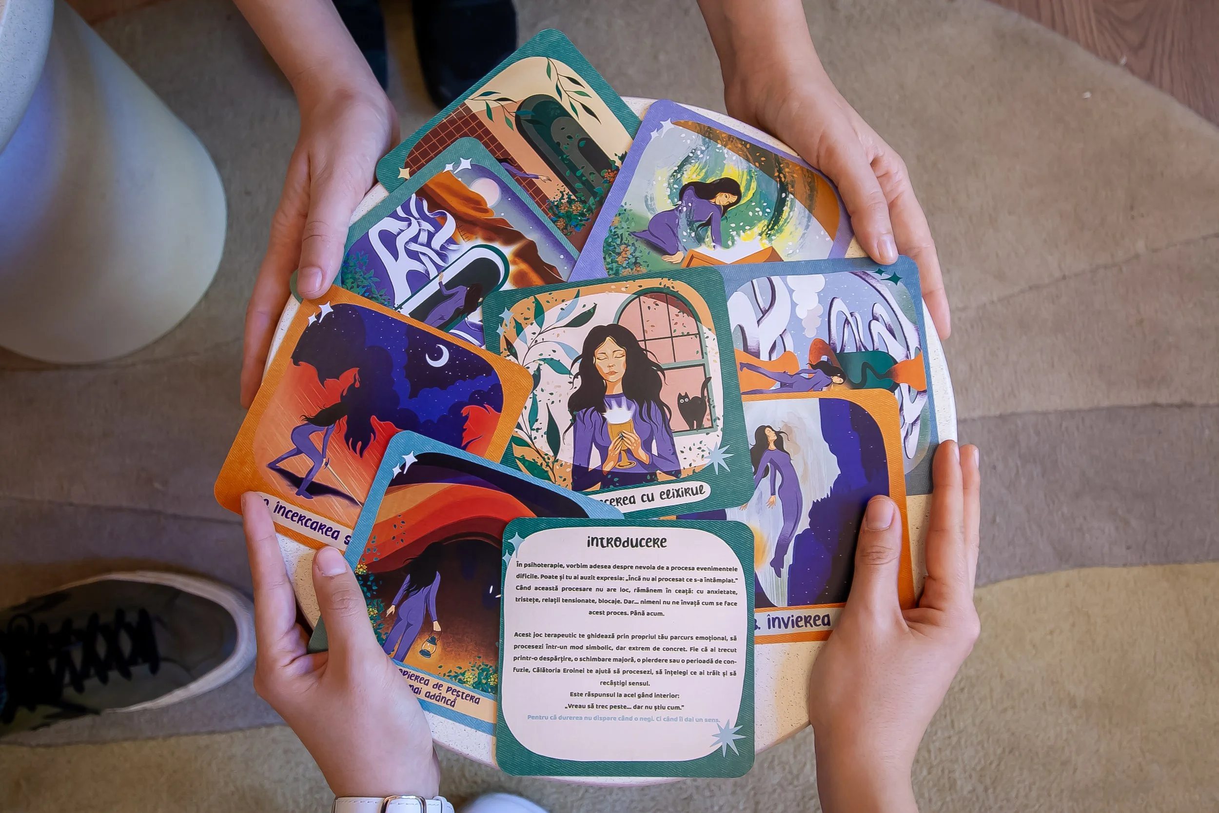 Hands holding open illustrated card deck with colorful fantasy-themed images of women, nature, and abstract scenes. - Carduri Calatoria Eroinei